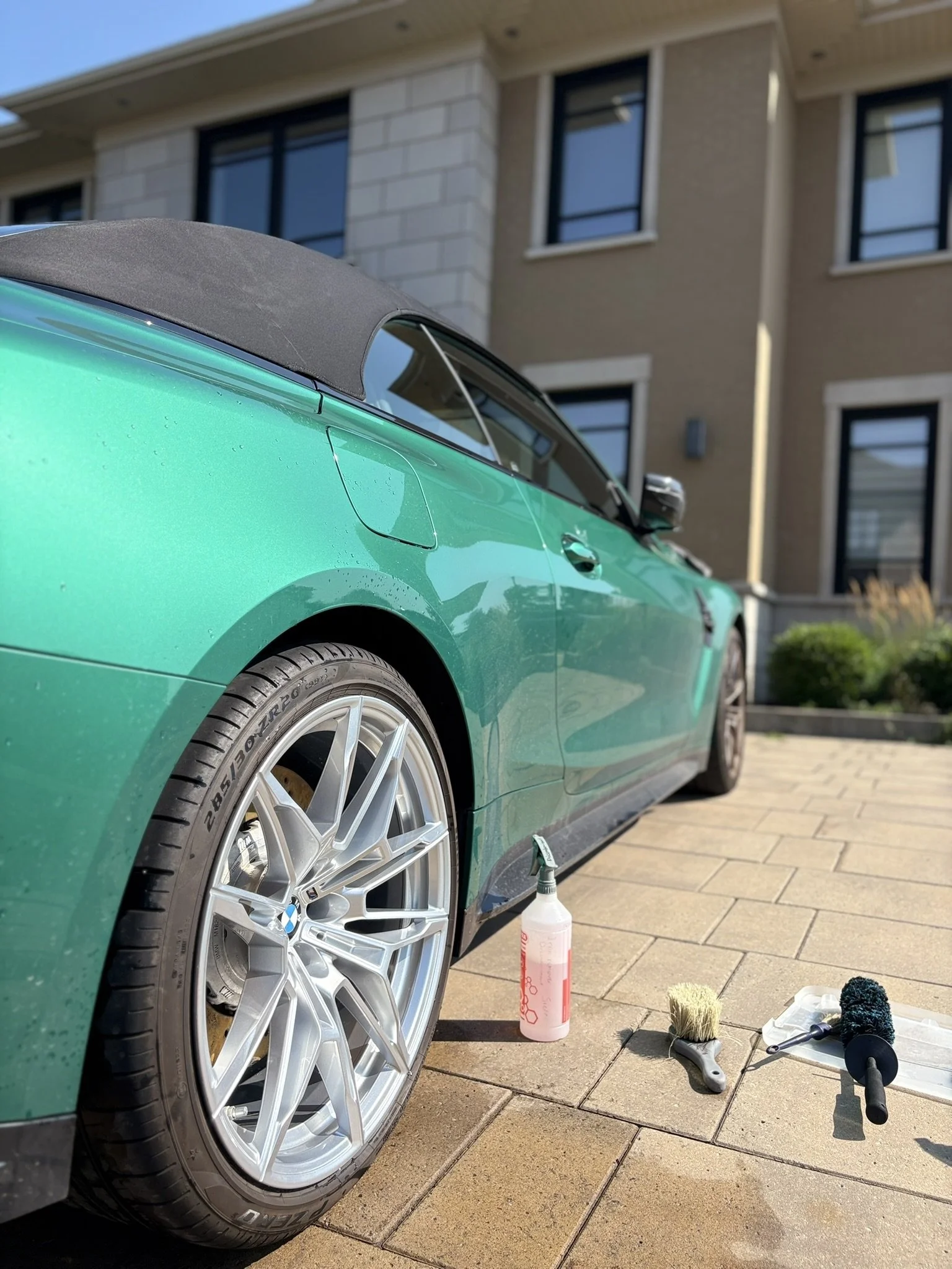 Voiture verte de marque BMW avec toit en tissu noir, en cours de nettoyage ou de polissage devant une maison. Des outils de nettoyage, dont une bouteille de spray, une brosse en crin et une brosse en mousse sont posés sur le sol en pavés.