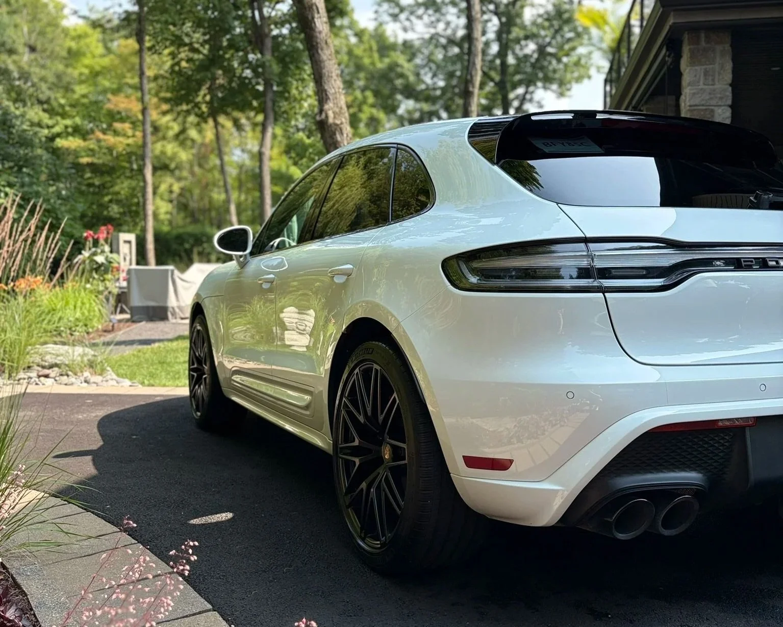 Voiture blanche de luxe Porsche Macan dans une allée de jardin ensoleillé avec arbres et plantes vertes.