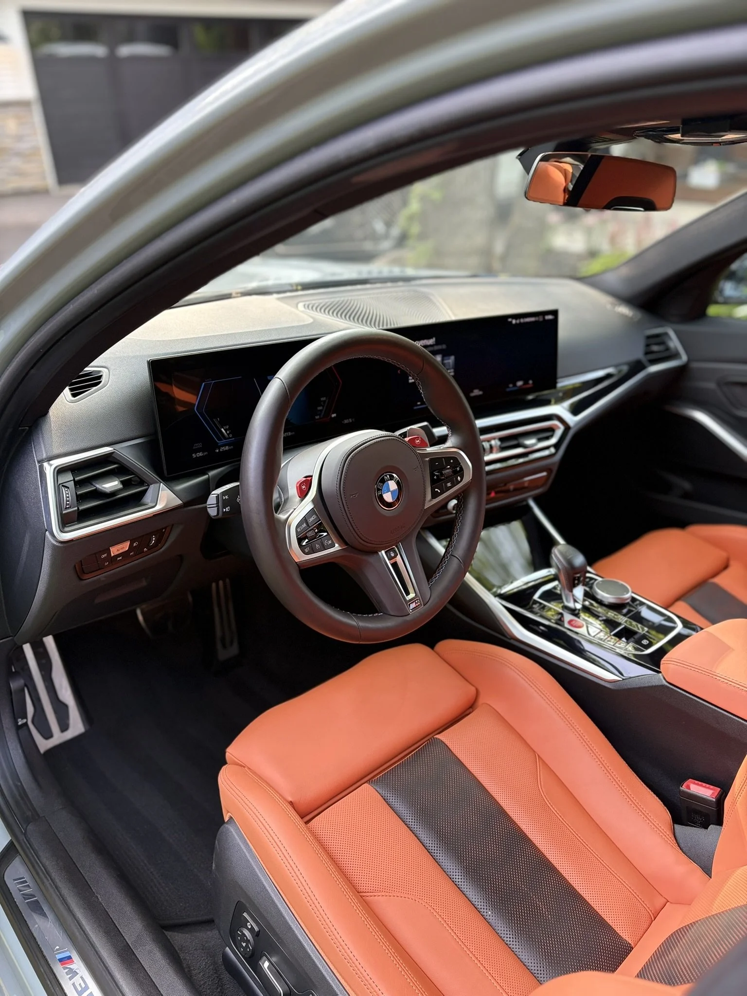 Intérieur d'une voiture BMW avec tableau de bord numérique, volant en cuir avec logo BMW, sièges en cuir orange, et centre de commande avec levier de vitesse et contrôleurs.