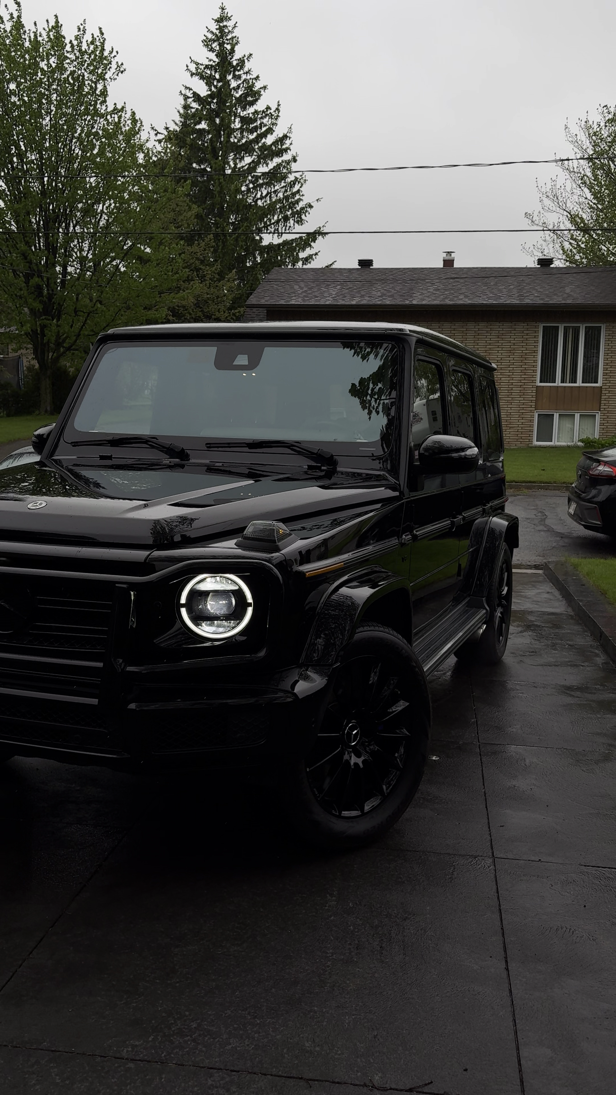 Voiture noire de style SUV avec phares allumés, stationnée sur un parking en extérieur par temps pluvieux devant une maison avec un toit en pente, entourée d'arbres et d'un ciel nuageux.