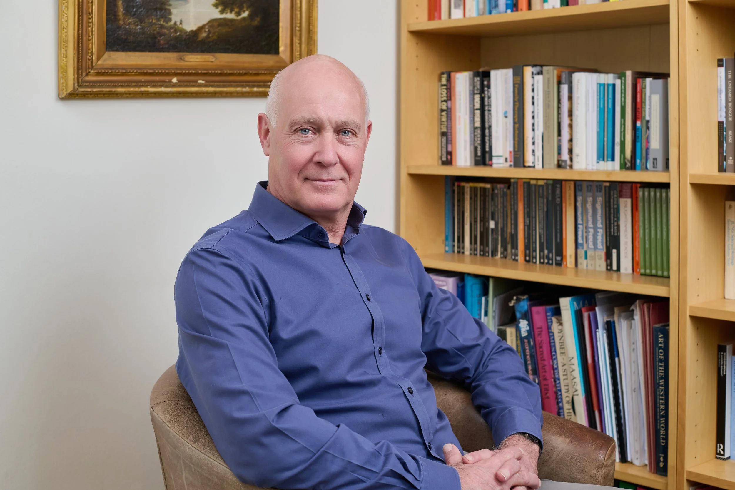 Dr William Hopkins wearing a blue button-up shirt, sitting in a chair with crossed hands, in front of a bookshelf filled with books.