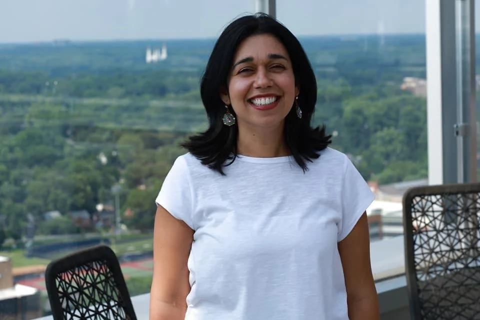 A woman with dark hair smiling in front of a window with a view of green trees and a distant white structure.