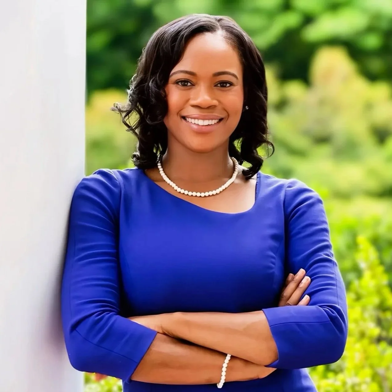 A woman in a blue dress and pearl necklace standing outdoors with her arms crossed, smiling at the camera, with greenery in the background.