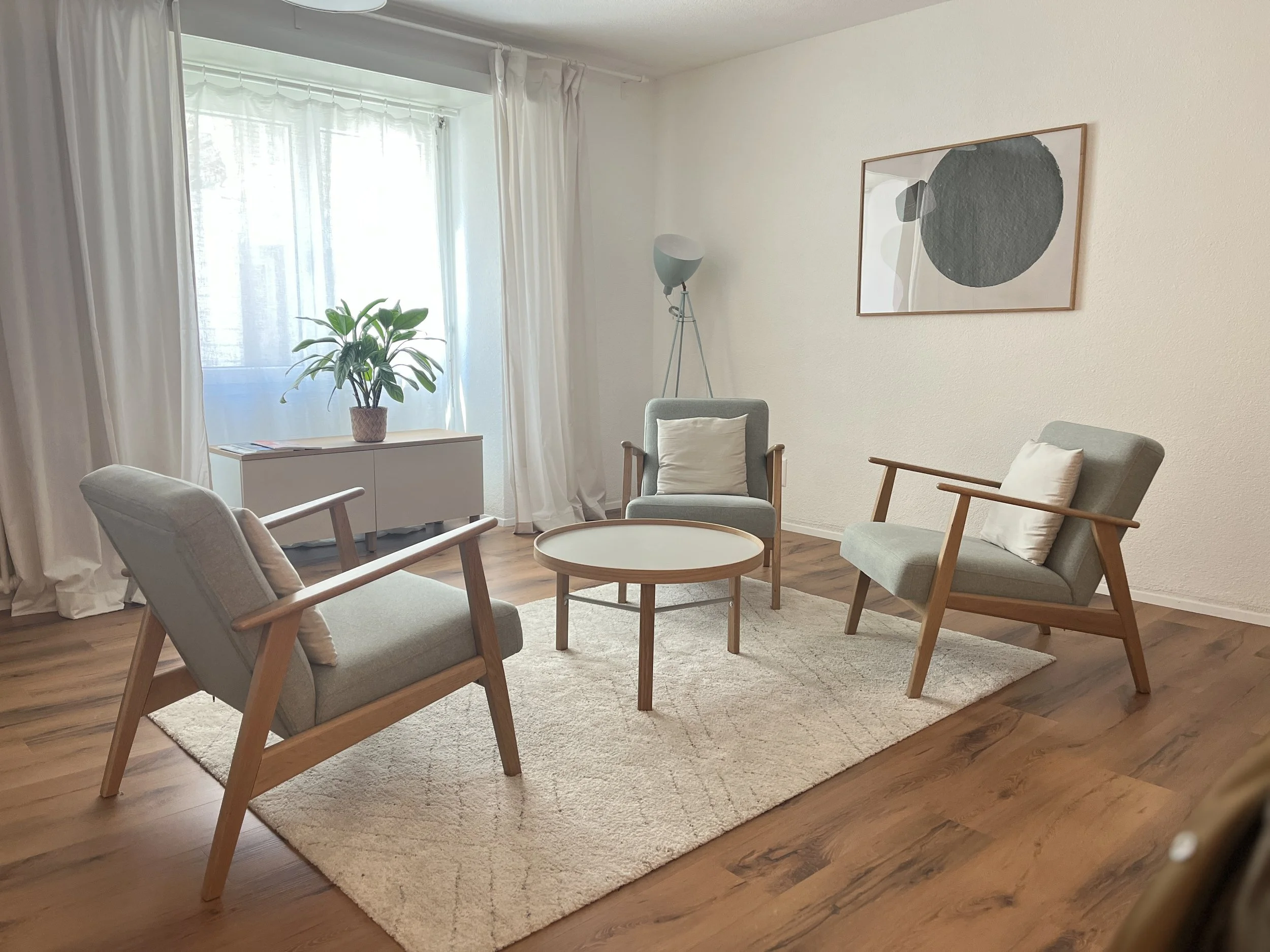 Ein helles Wohnzimmer mit vier modernen Stühlen, einem kleinen runden Tisch, einer weißen Kommode mit Topf-Pflanze, einem stehendem Lampen und einem Wandbild mit abstraktem schwarzen und grauen Kreis. Praxis für psychologische Beratung für Frauen