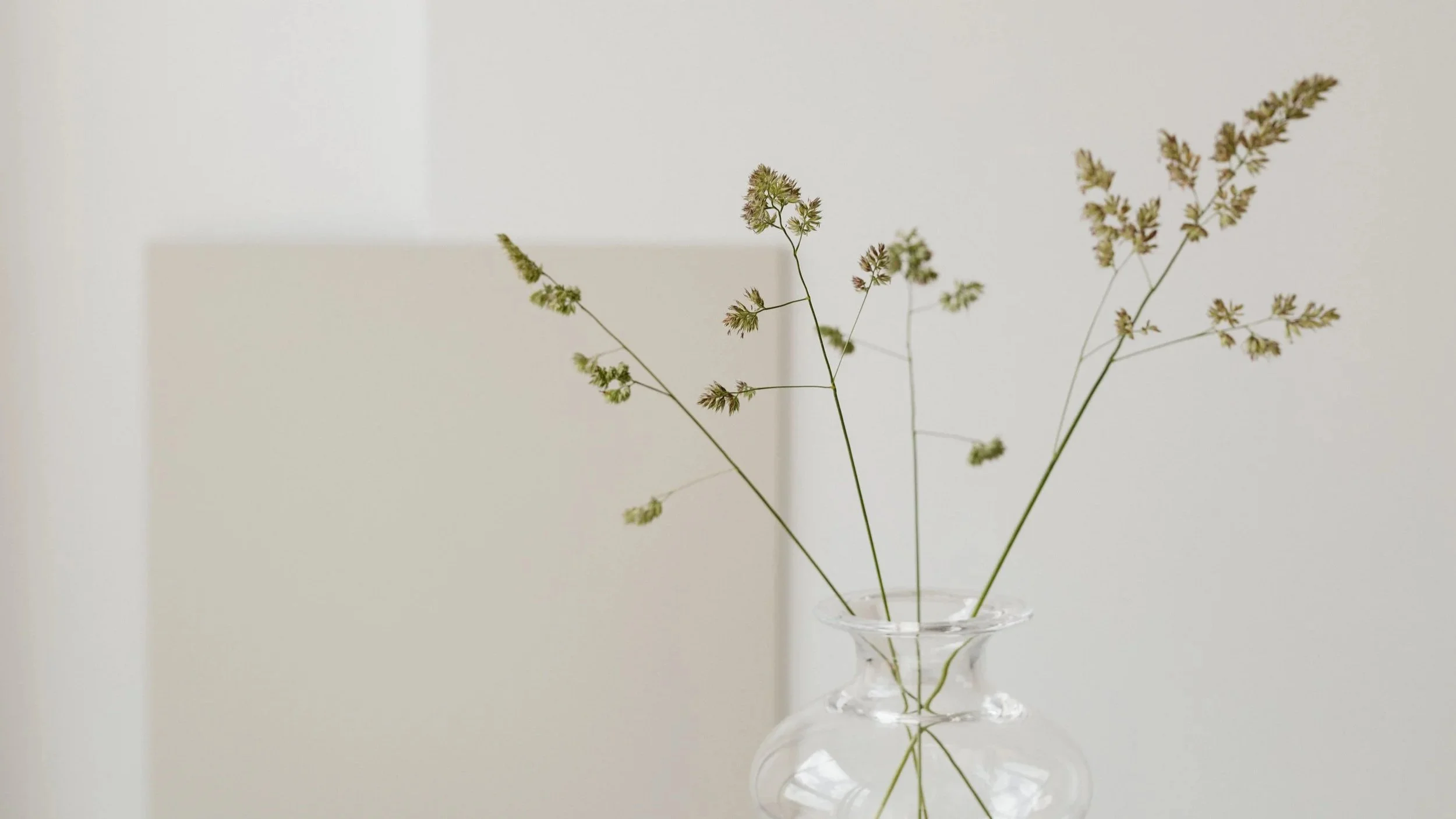 Eine transparente Vase mit grünen Zweigen und Blüten vor einem hellen, neutralen Hintergrund.