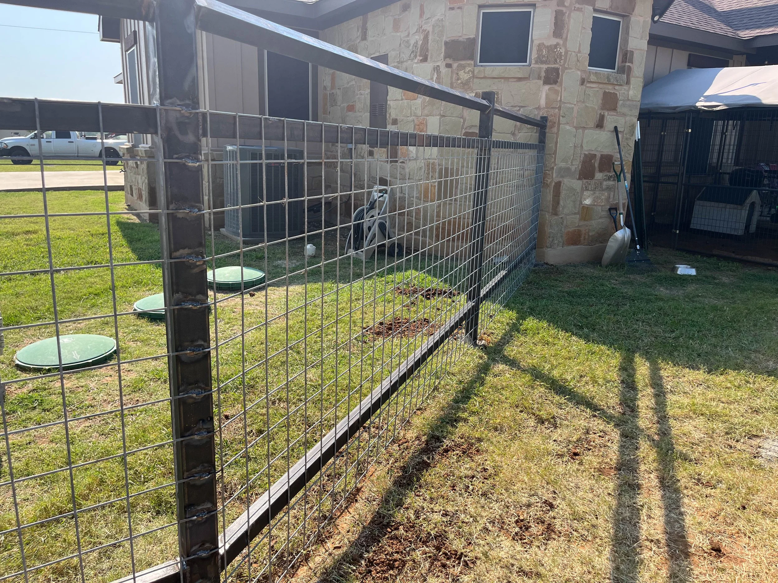 View of a backyard yard with a metal fence, a stone house facade, and gardening tools leaning against the house, under a bright clear sky.