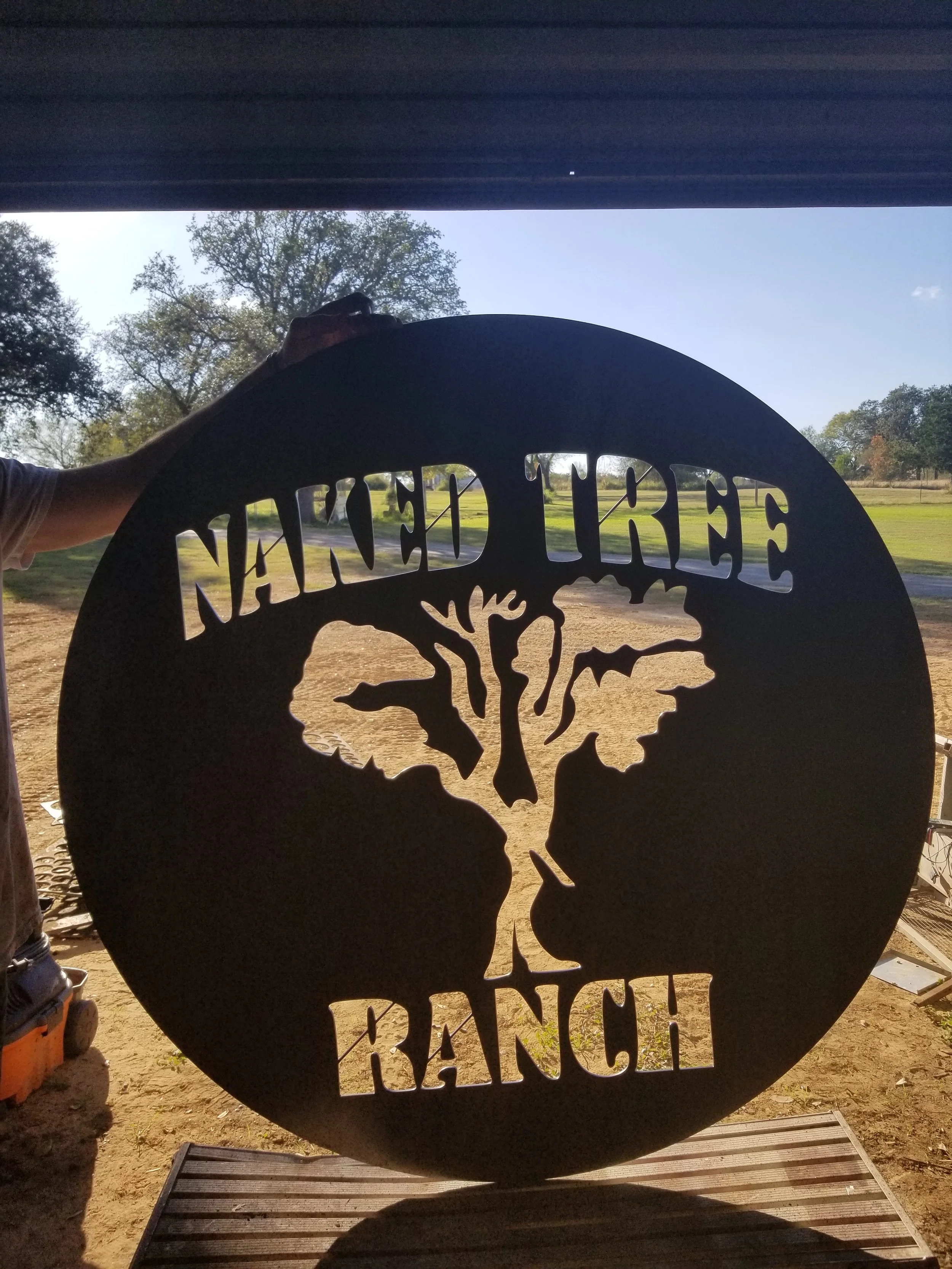 A circular metal sign with the design of a skull and the words 'Learned Tree GANG!' cut out of it, placed outdoors with trees and a grassy field in the background.