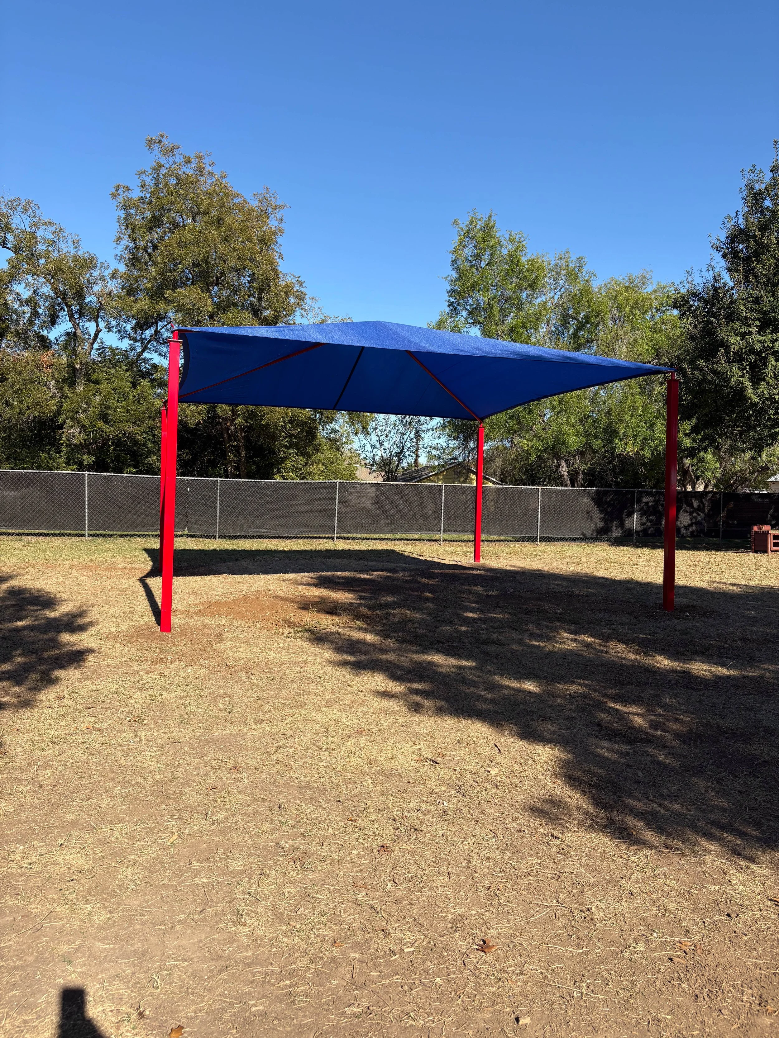 A blue shade structure with red poles is set up in an outdoor area with dirt ground, surrounded by trees and a chain-link fence, under a clear blue sky.