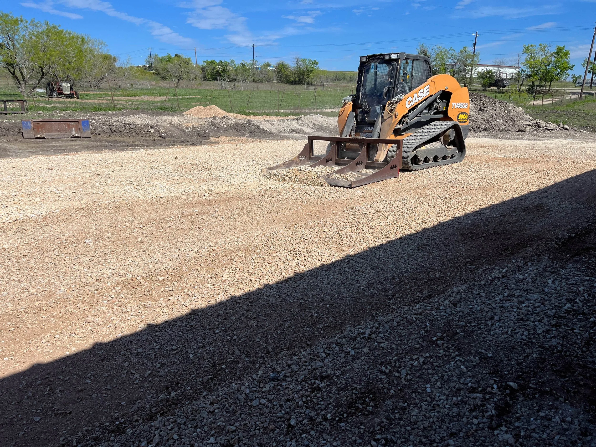 A small construction loader sits on dirt and gravel in an outdoor area with trees, grass, and utility poles in the background under a blue sky.
