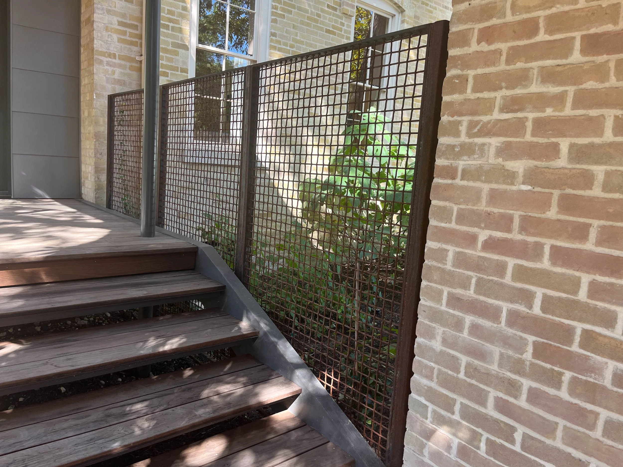 Exterior view of a residential building with brick and siding walls, a wooden deck, metal post, and a metal fence with a plant growing behind it. A window with white trim is visible on the house.