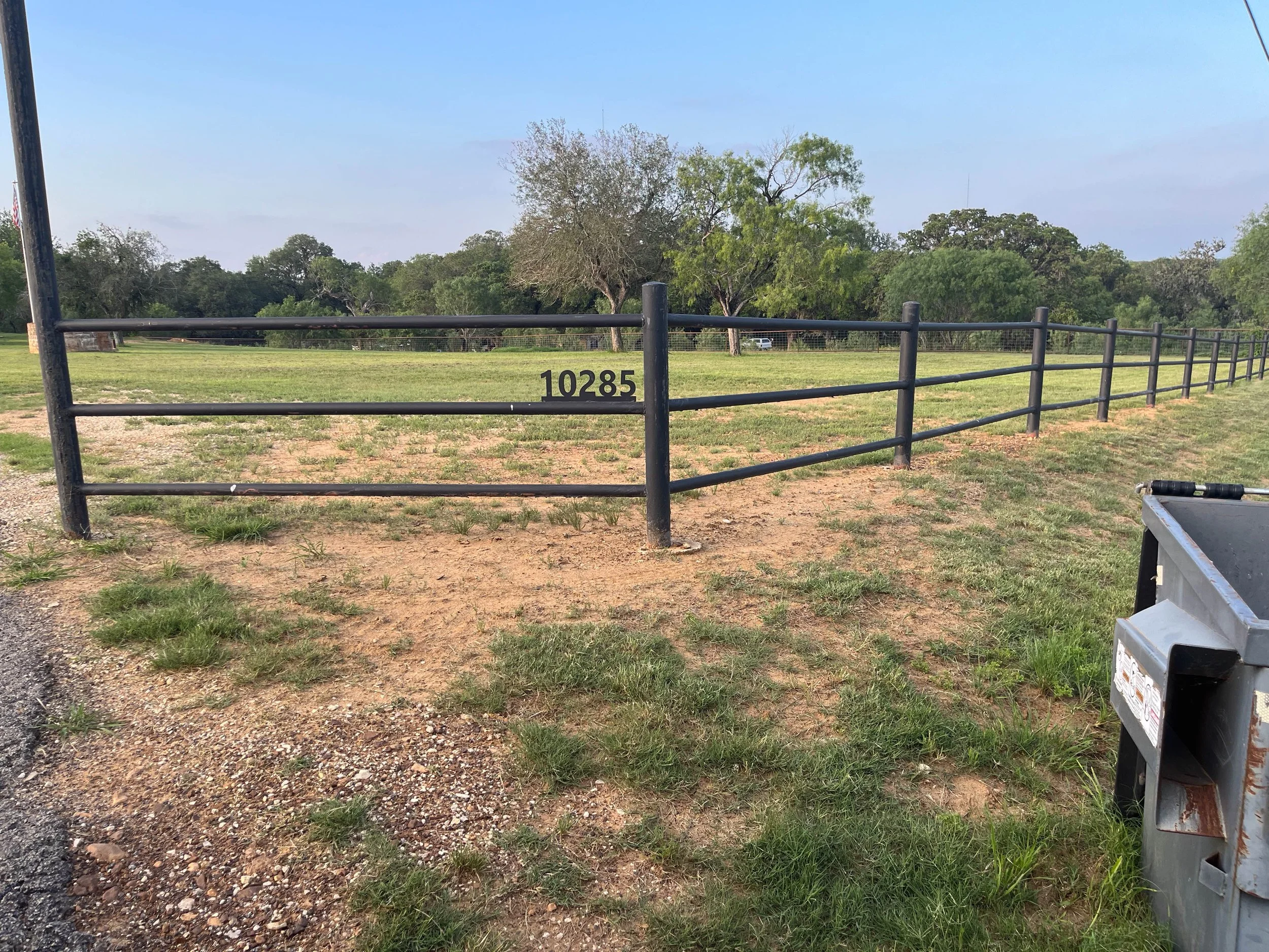 A black metal fence with a small gate and a number 10285 near a grassy field with trees in the background and a partly cloudy sky.