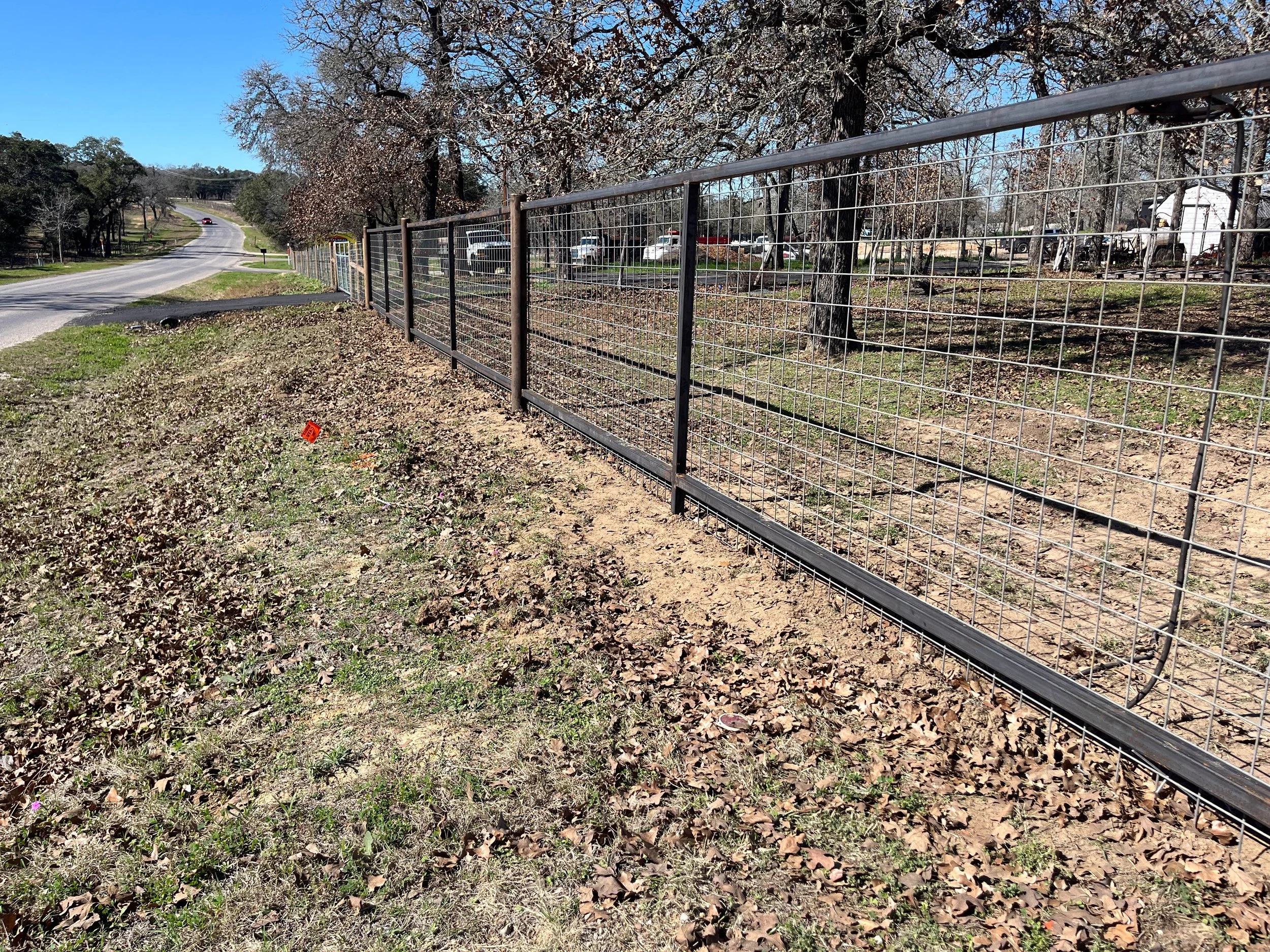 A metal fence running along a grassy area with trees, some with leaves, across a street from a parking lot with multiple cars and a trailer, under a clear blue sky.