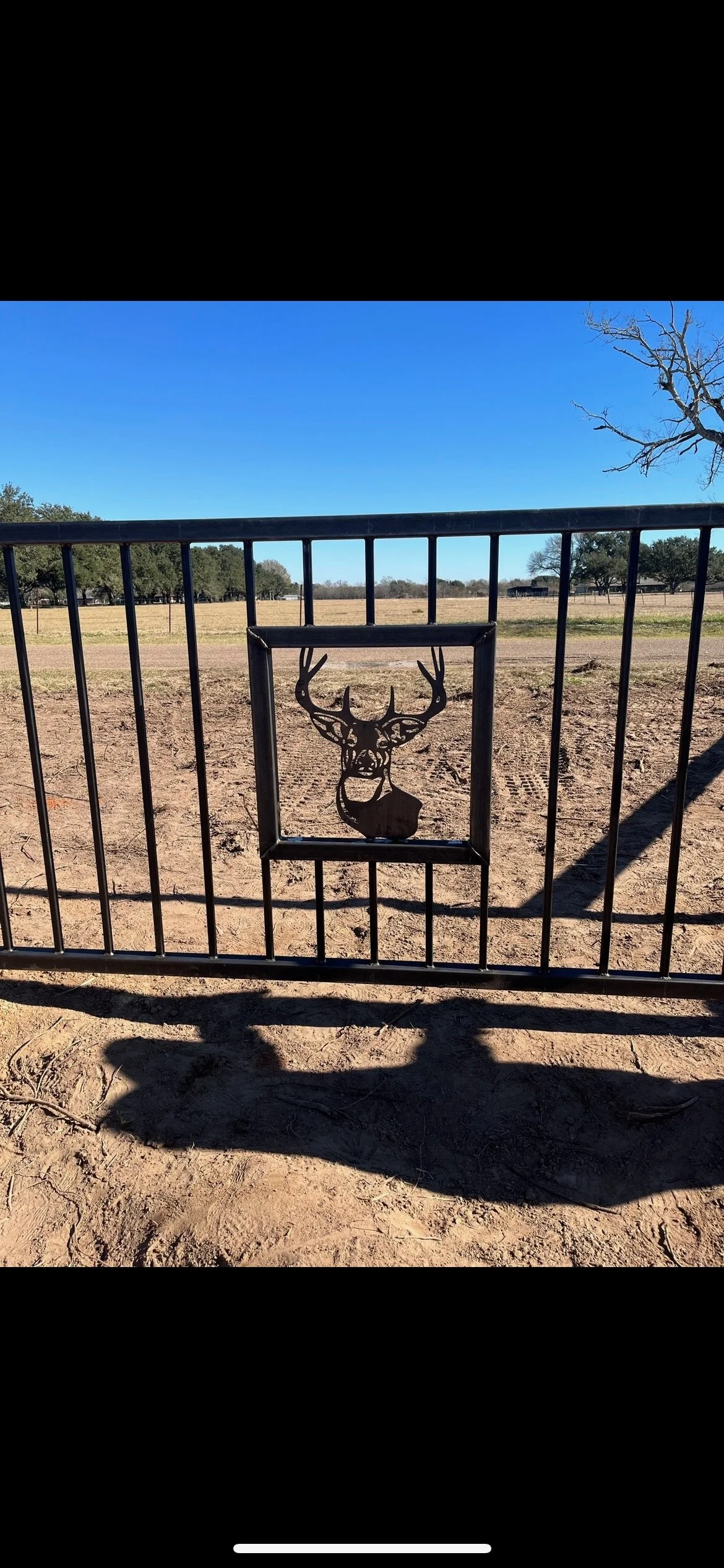 Black metal gate with a cutout silhouette of a deer head with antlers, set against a rural landscape with open field, trees, and a clear blue sky.