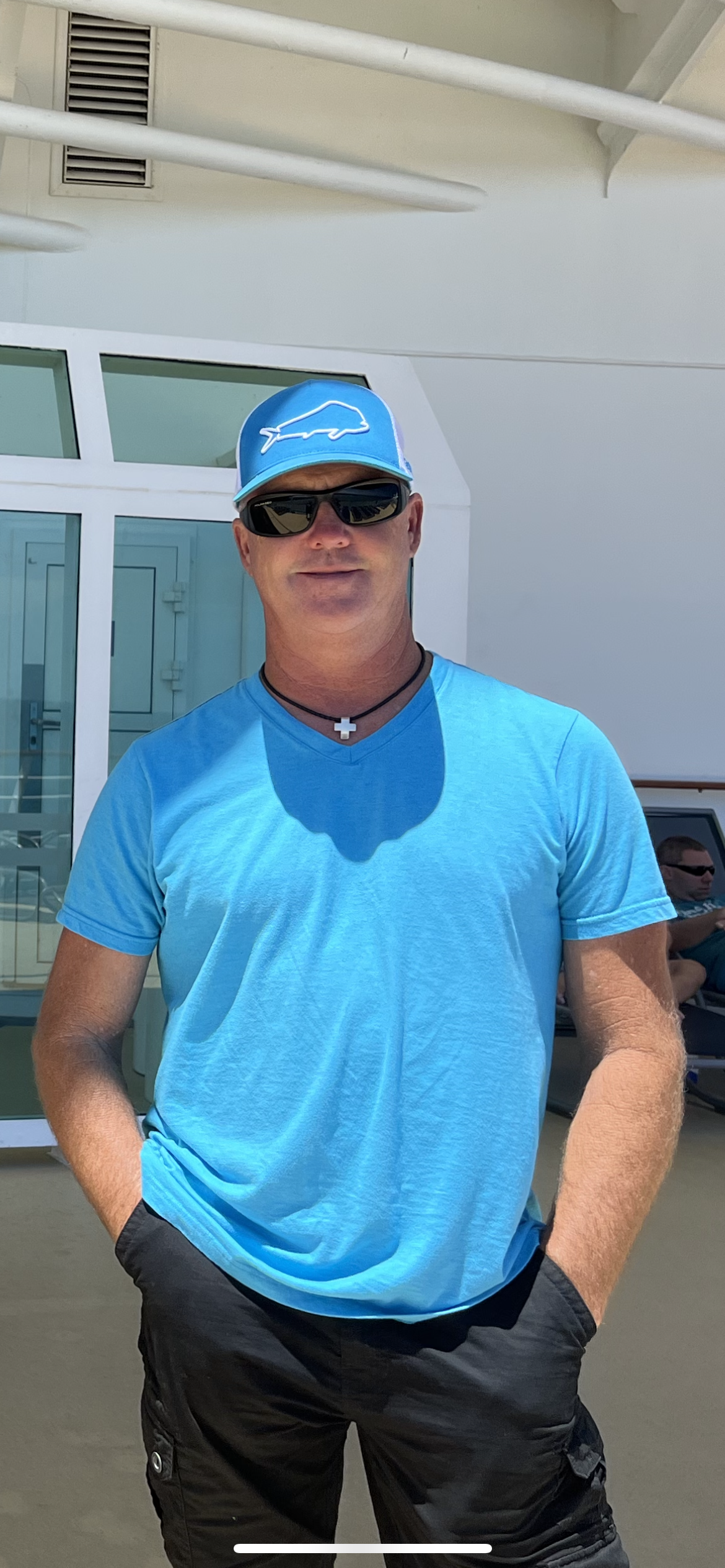 A man wearing a blue t-shirt, black cargo shorts, sunglasses, a blue cap with a logo, and a necklace with a cross pendant, standing outdoors near a building with a glass door and window.