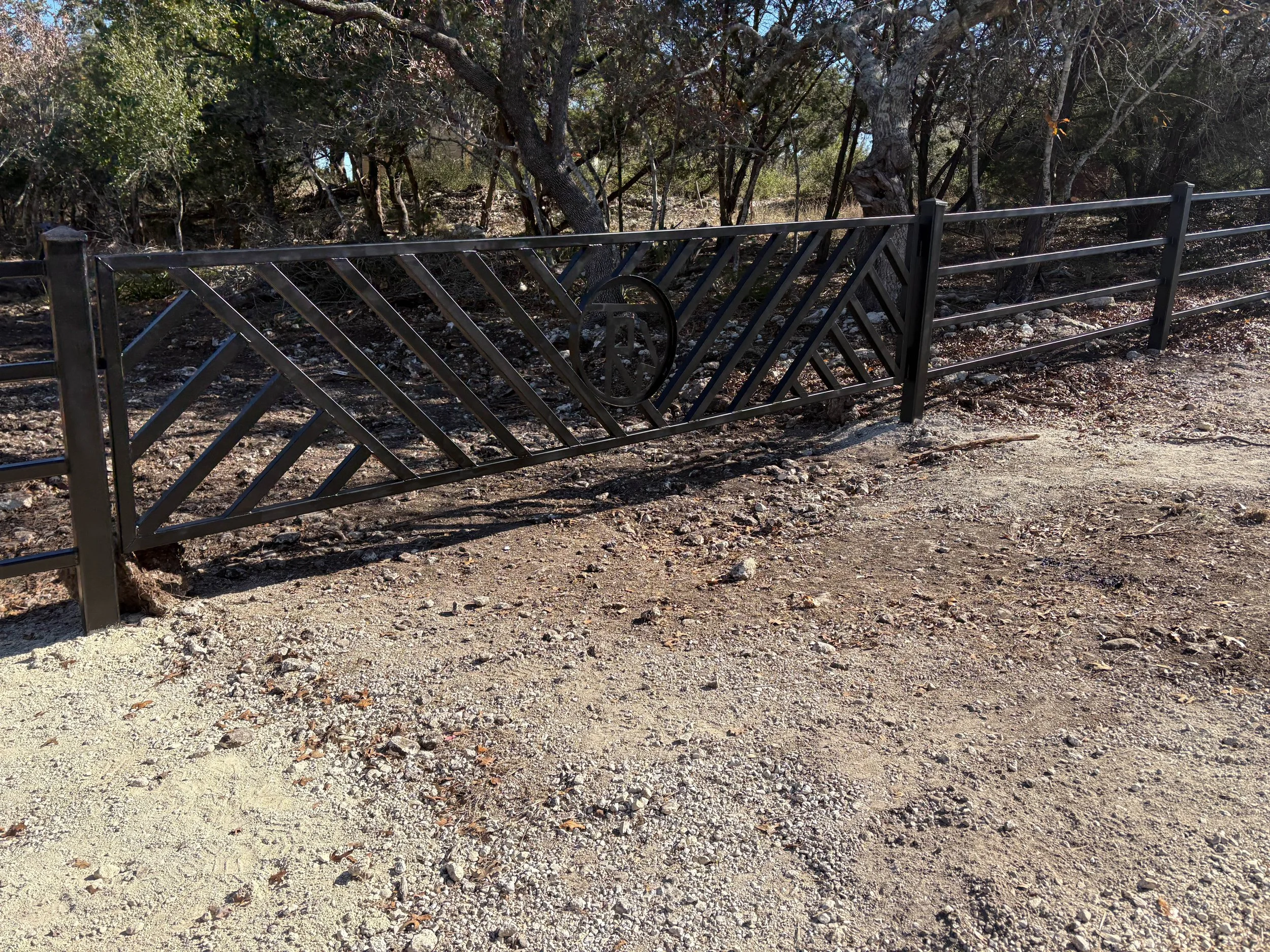 A black metal gate with a diagonal pattern blocks the entrance to a wooded area with trees and bushes.