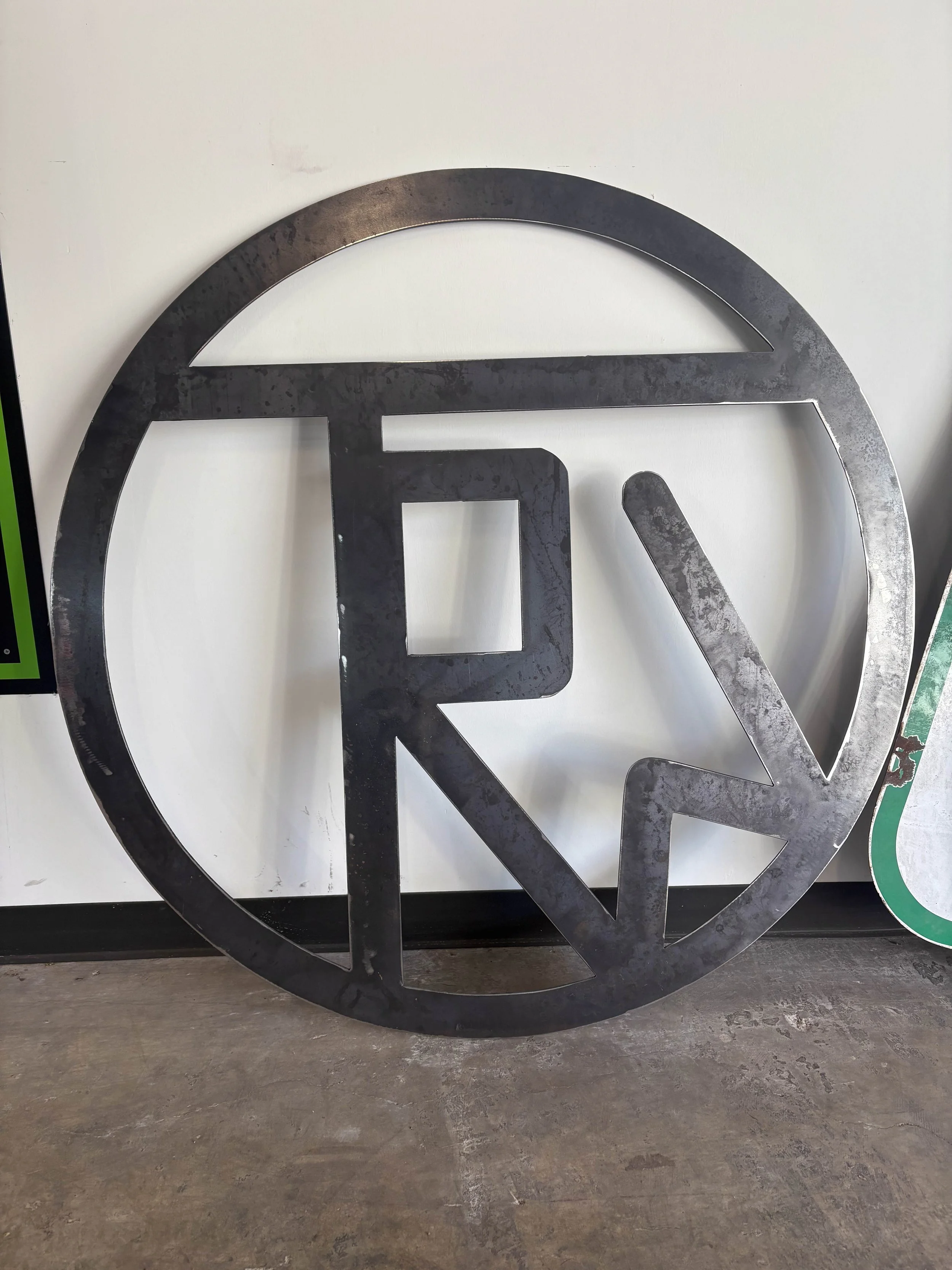 Large metallic logo with circular and square elements resting on a concrete floor, with a white wall in the background.
