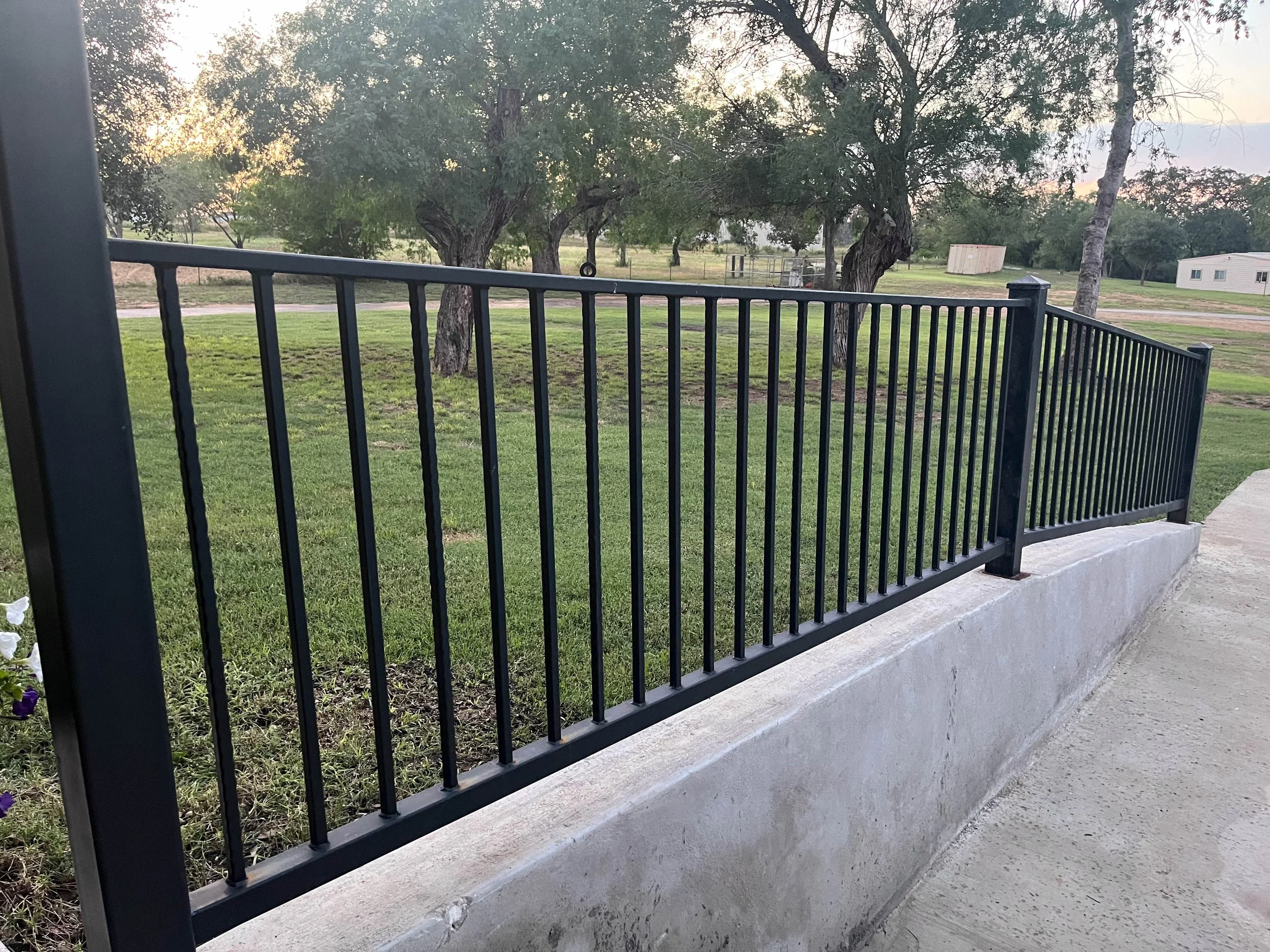 View of a backyard with a black metal fence, green grass, several trees, and a few small structures in the distance during sunset.