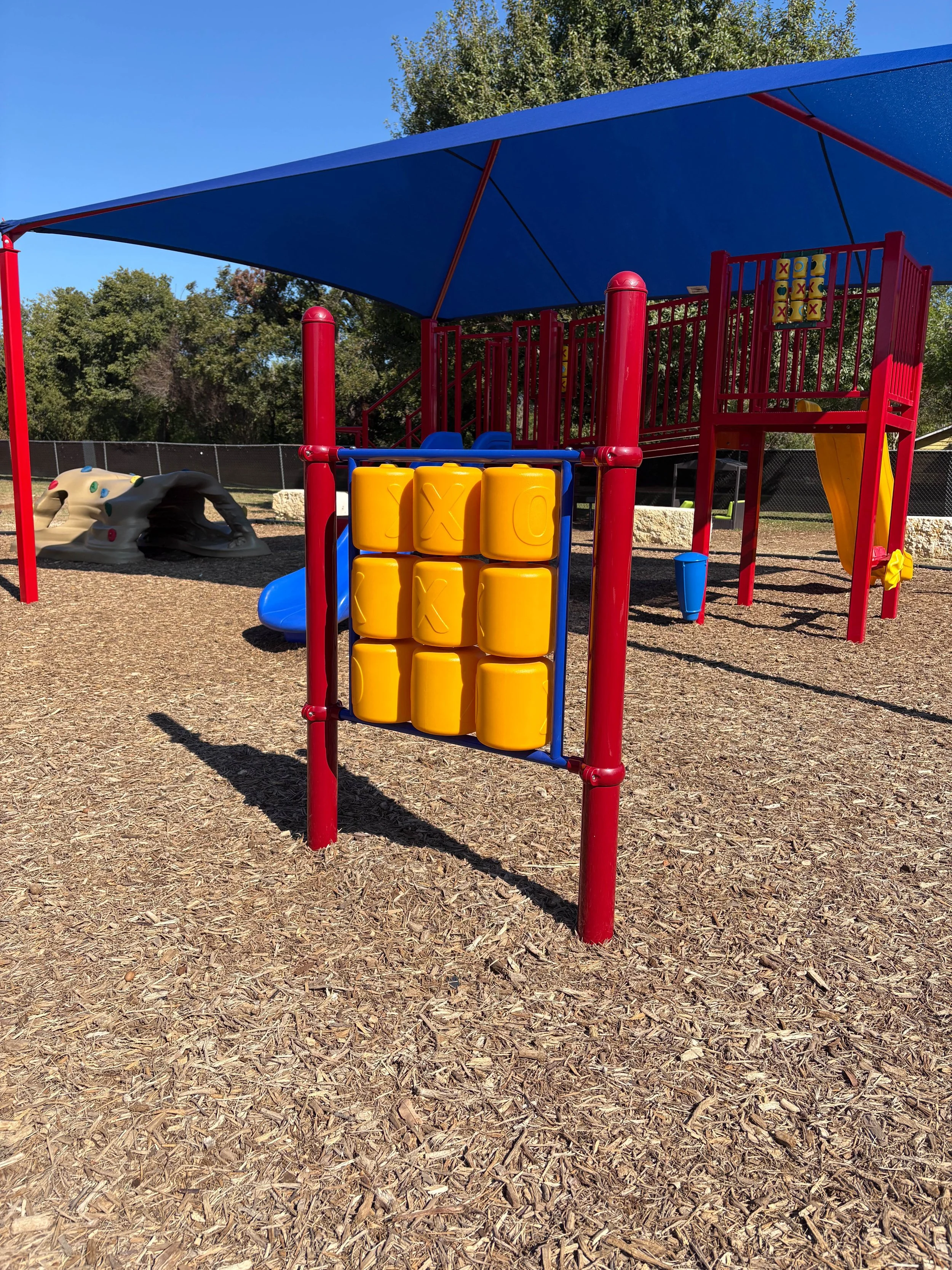 A colorful playground with a blue shade canopy, red supports, and various climbing and sliding structures, all on a wood chip ground surface.