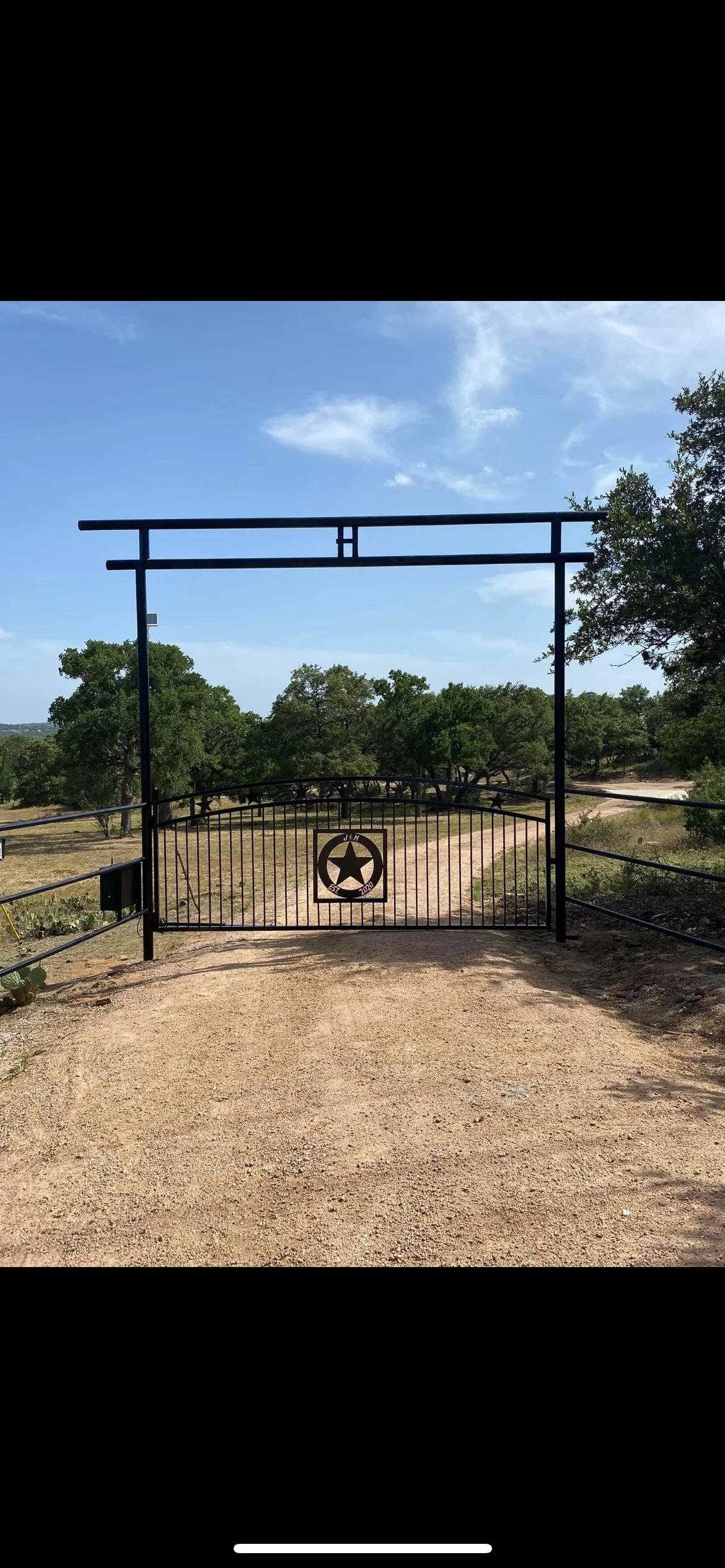 A black metal gate with a star emblem and the letters 'J M' on a dirt road in a rural landscape with trees and a partially cloudy sky.