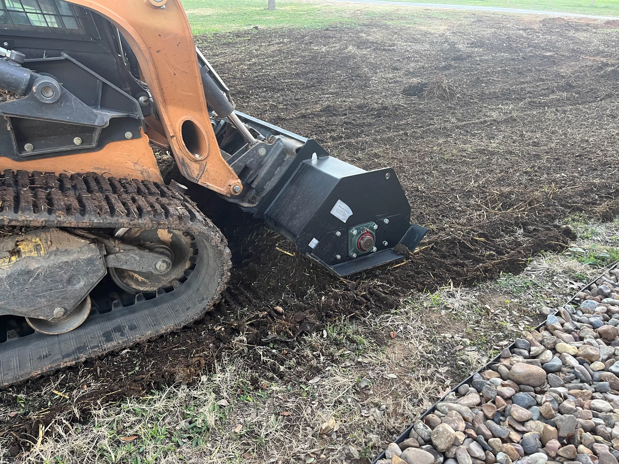 Close-up of a tracked bulldozer with black and orange equipment leveling a dirt field, with a grassy field in the background.