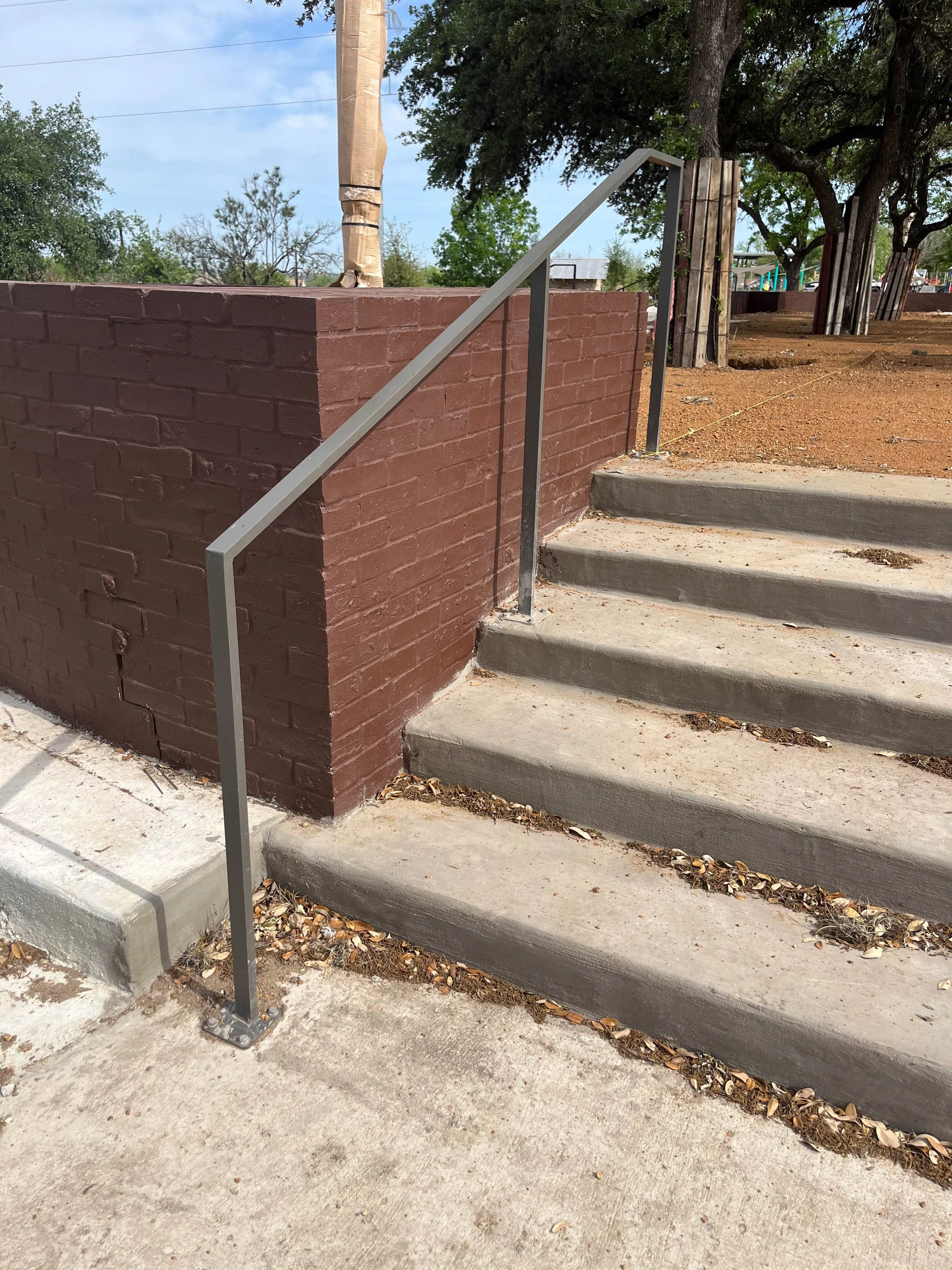 Concrete stairs next to a red brick wall with a metal handrail, outside in a park or playground area with trees and dirt ground.