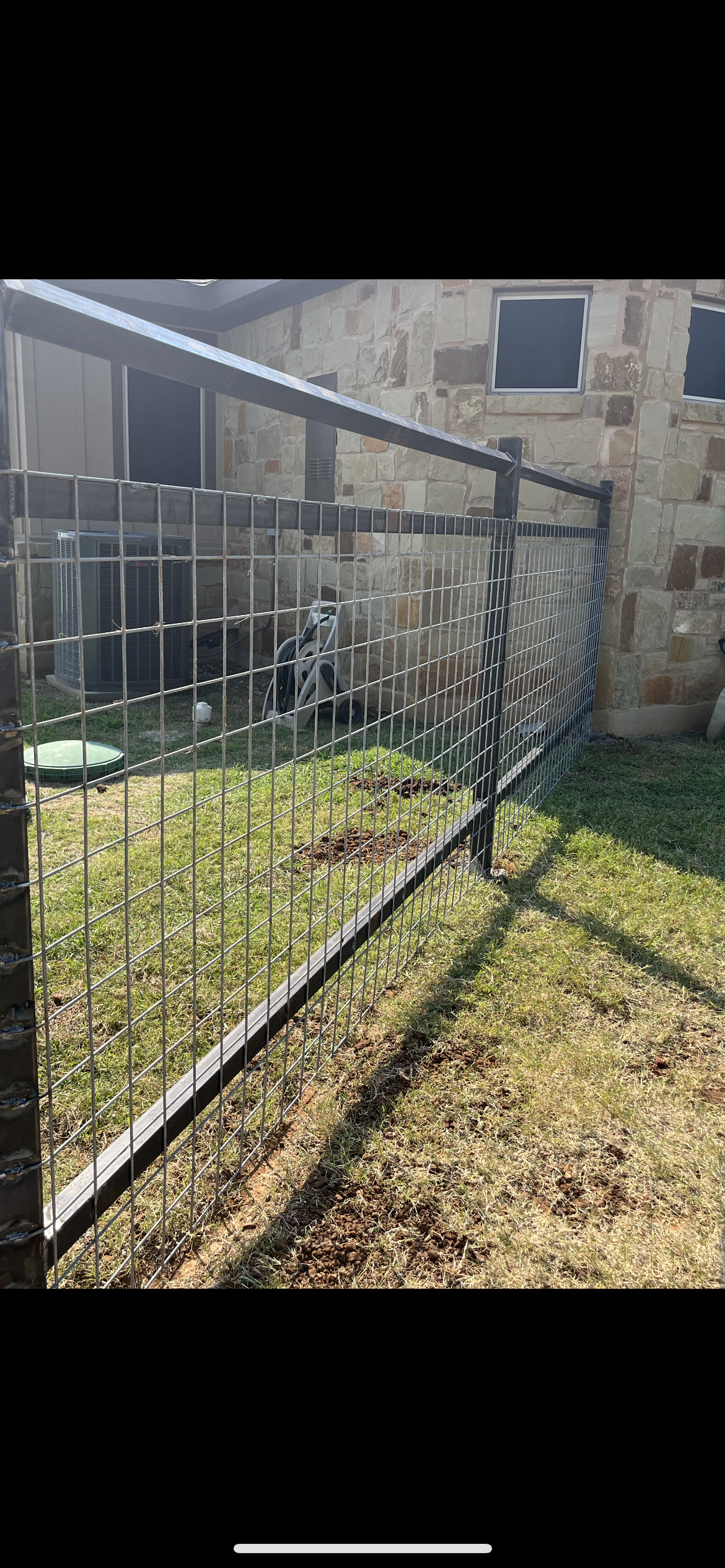 A black metal wire fence enclosing a small grassy yard with an air conditioning unit, a garden hose, a green lid, and some patches of disturbed earth, adjacent to a stone wall house.