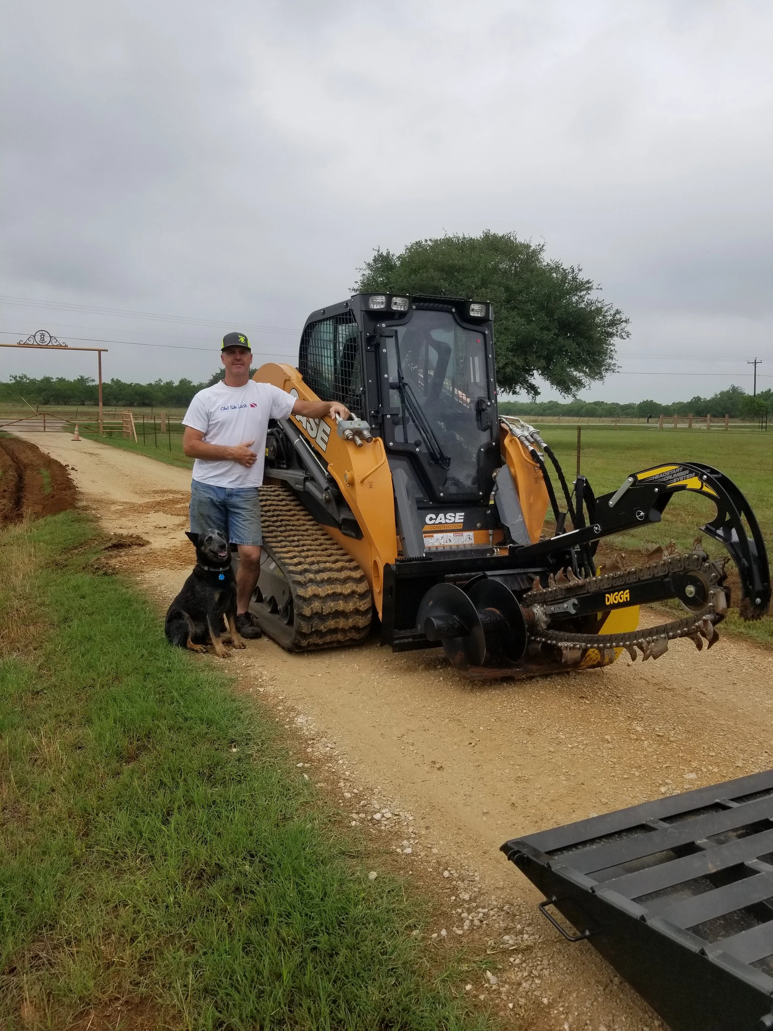 A man standing next to a small orange and black CASE mini excavator with a dog sitting nearby, on a dirt path in a rural area with trees and a cloudy sky in the background.