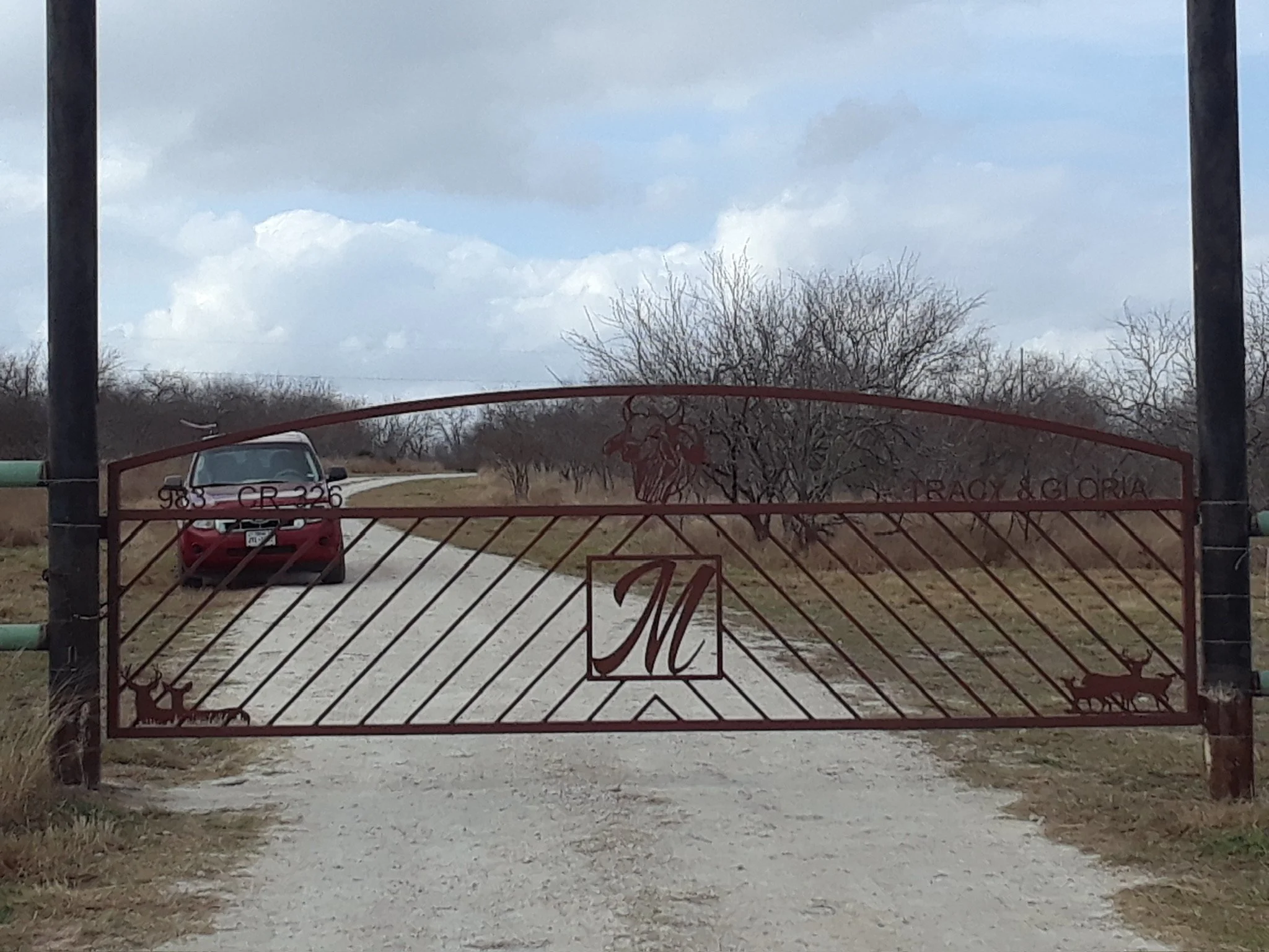 A red metal gate with the words 'GRACIA GLORIA' is open, revealing a driveway with a red car parked under a cloudy sky and leafless trees in the background.