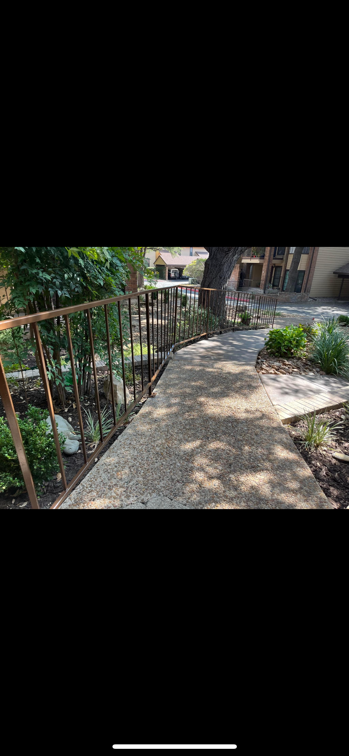 Curved sidewalk with a metal railing on the left and plants on both sides, with trees and residential buildings in the background.