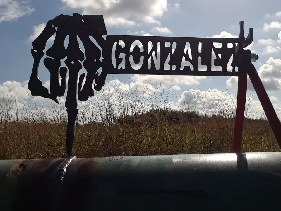 Silhouette of a skeletal horse head and the text 'GONZALEZ' on a metal sign, set against a cloudy sky with grassy field in the background.