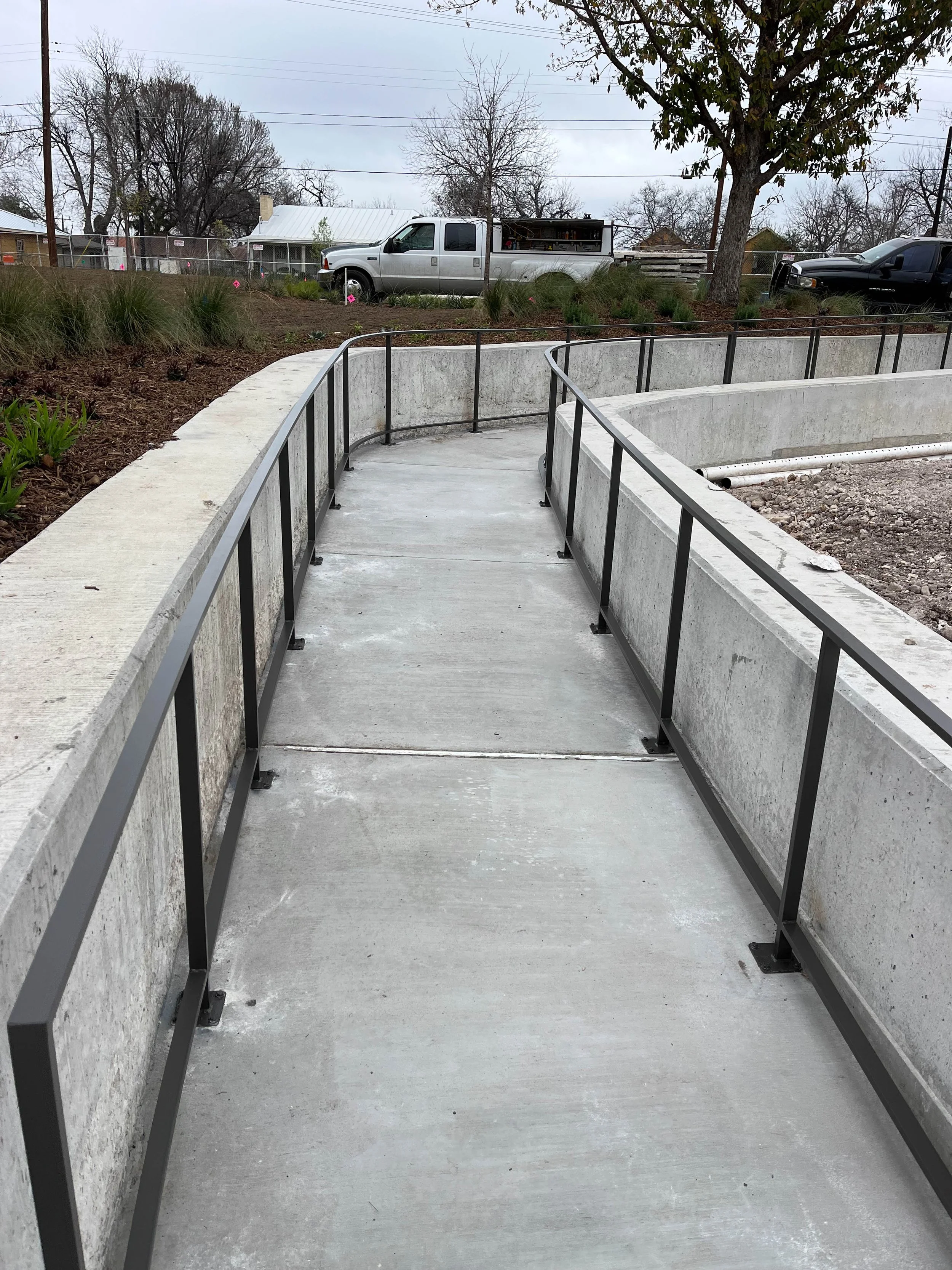 Concrete walkway with black metal handrails, leading to a landscaped area with bushes, trees, parked trucks, and a chain-link fence in the background.