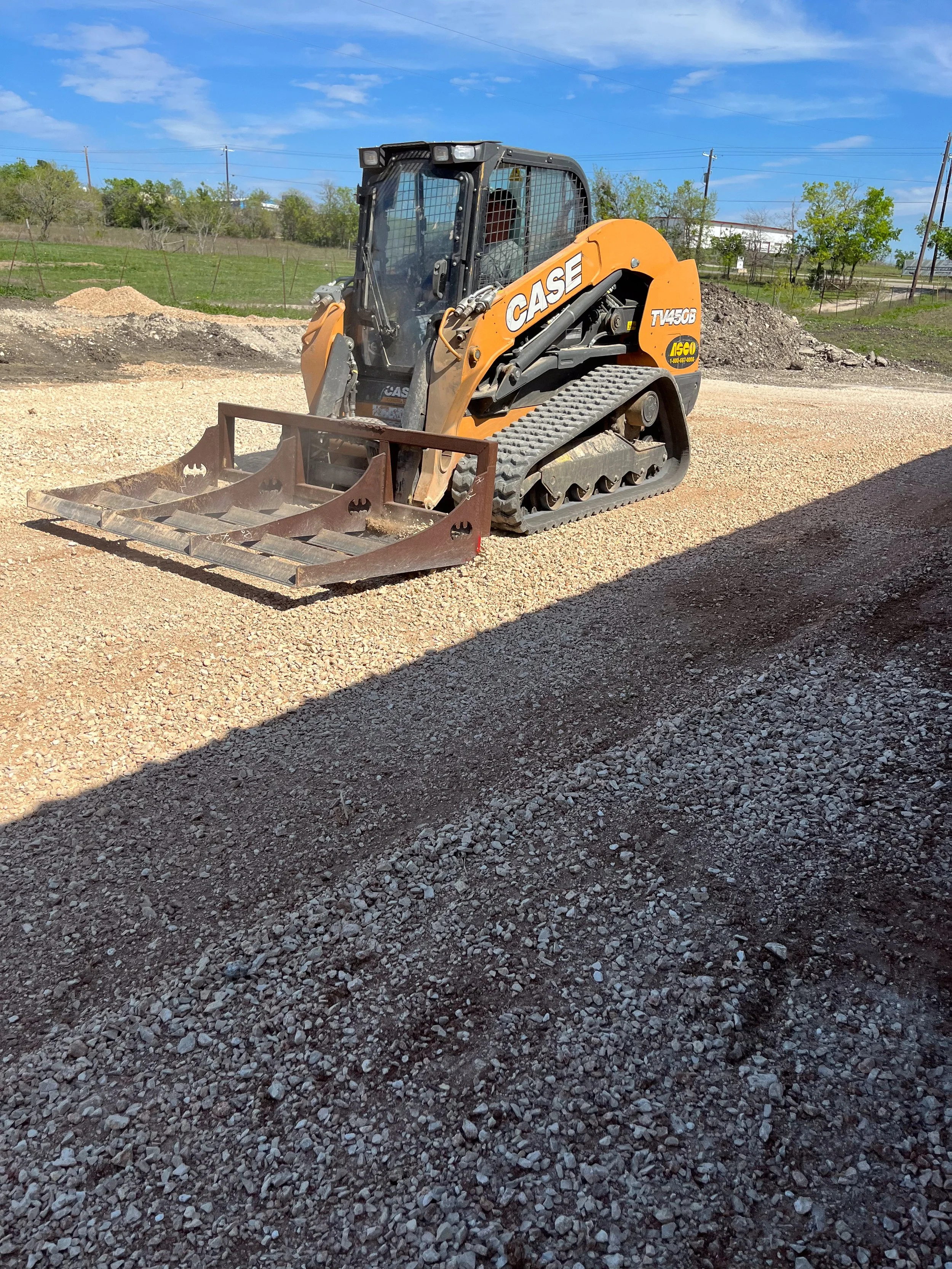 A small orange and black CASE mini excavator on a dirt and gravel construction site with piles of dirt, a fence, trees, and a blue sky in the background.