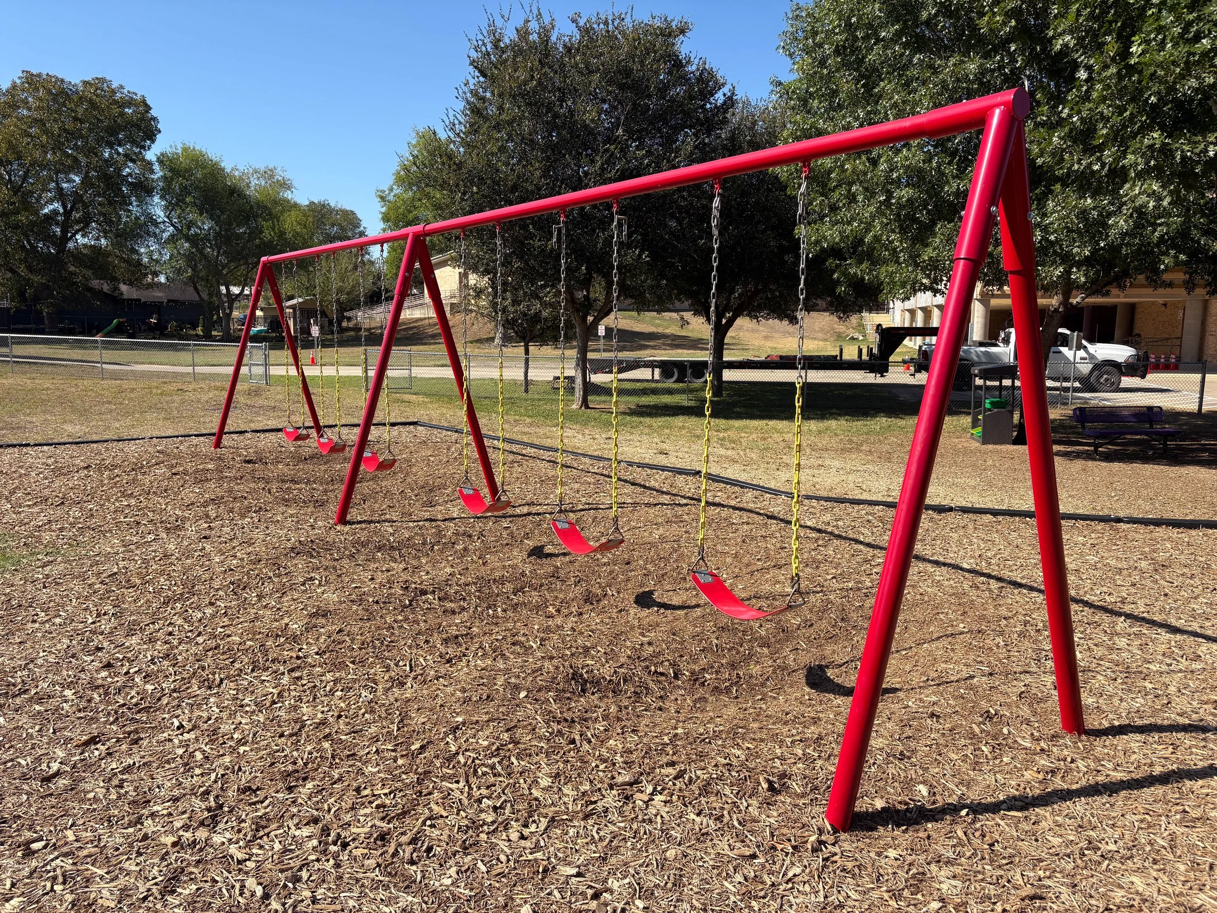 A playground with a red swing set and four swings, surrounded by trees, a fence, and a parking lot with vehicles beneath a clear blue sky.