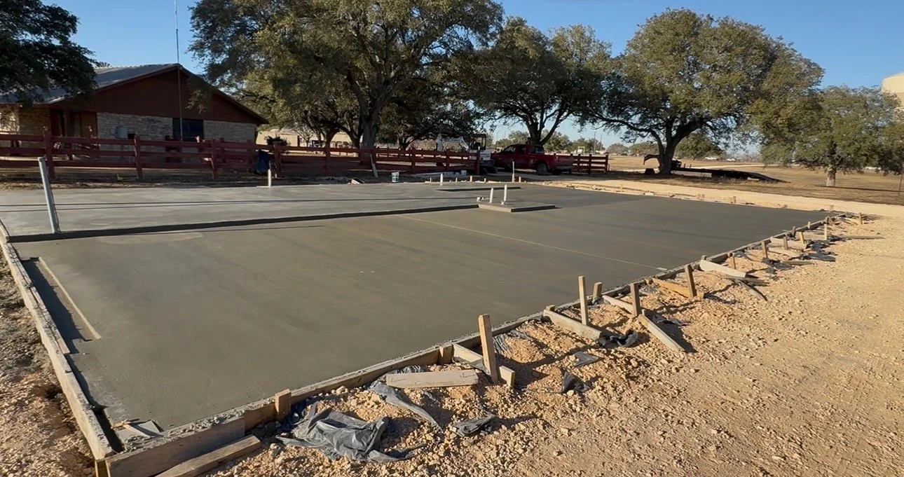 Freshly poured concrete on a construction site for a small sports court, with wooden framing and stakes, in a backyard with trees and a red fence.