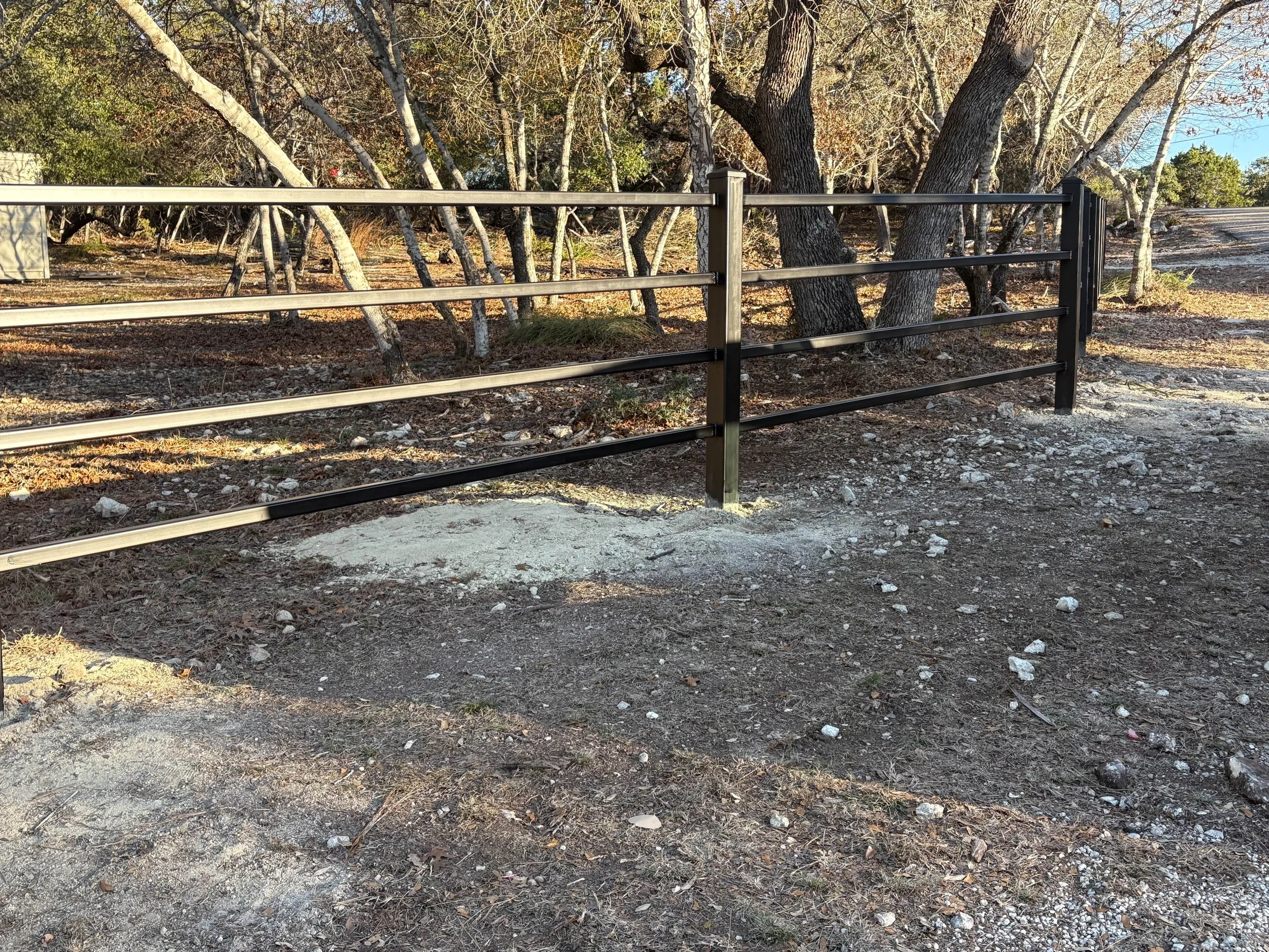 Image of a metal fence with trees and a dirt path behind it, sunlight illuminating the scene.