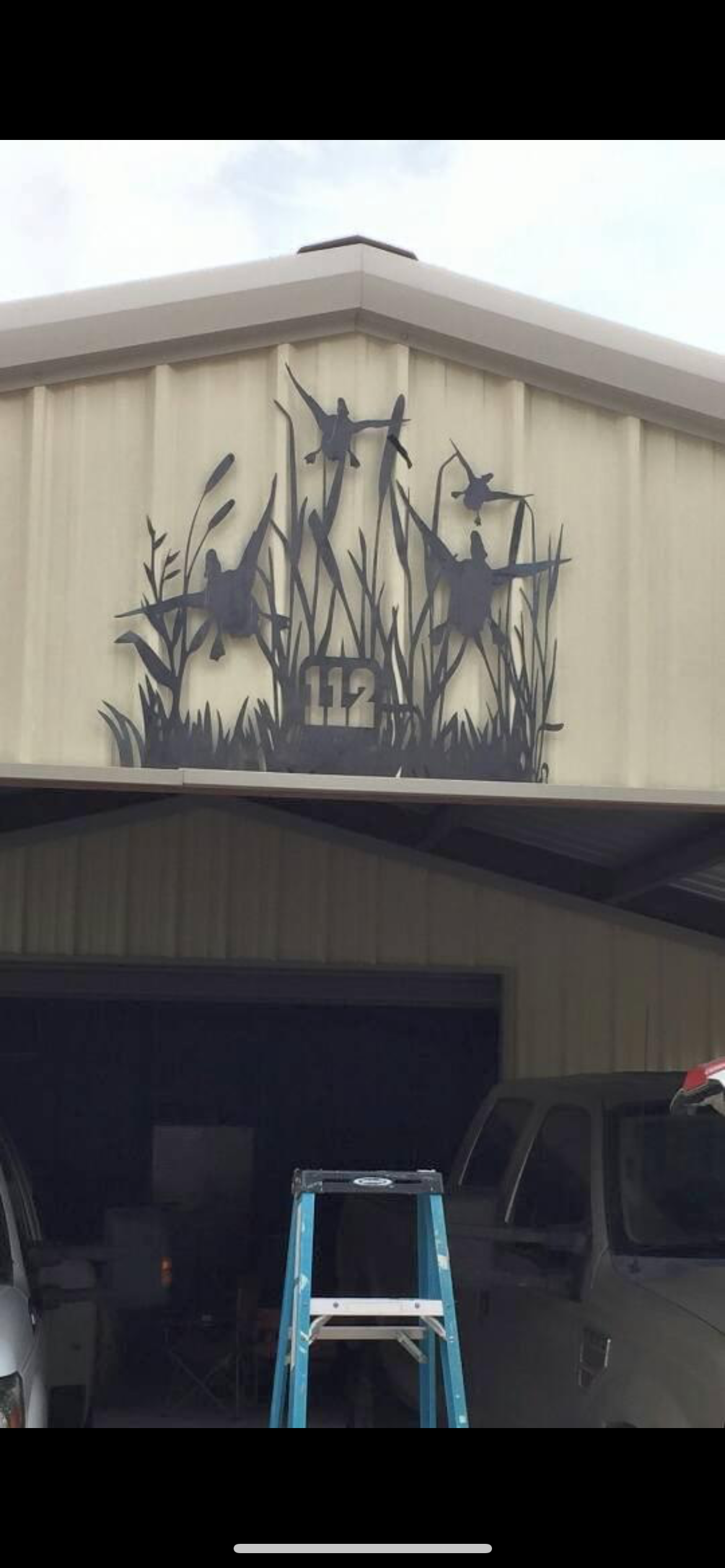 Metal silhouette of three birds perched and flying among reeds, with the number 112 below, mounted on the exterior wall of a building.