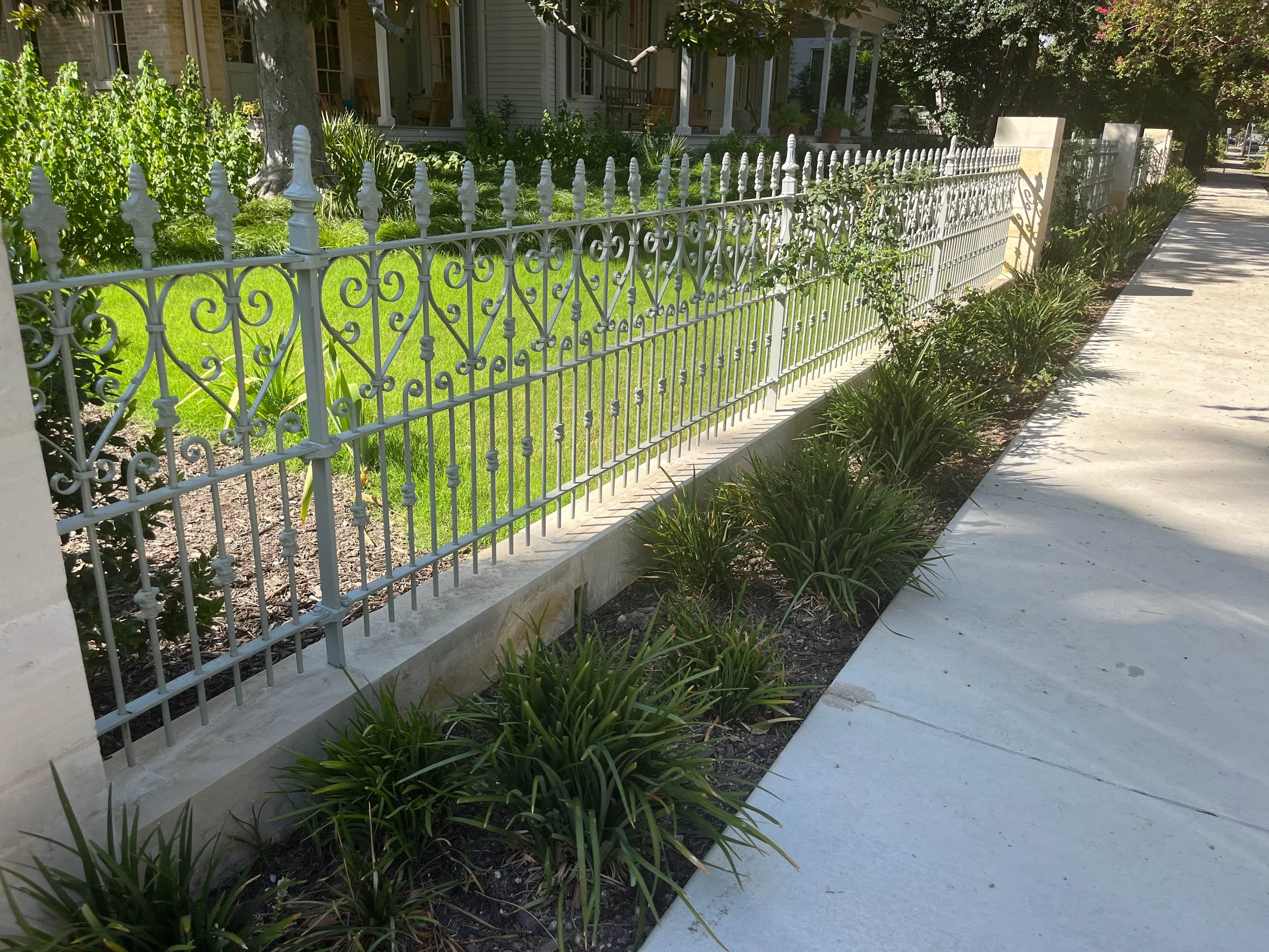 White decorative metal fence along the sidewalk in front of a house with a porch and garden, with green bushes and plants