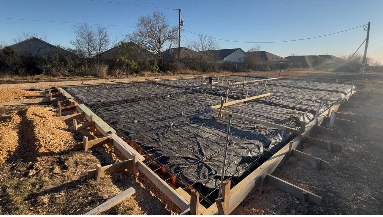Construction site with a foundation prepared for pouring concrete, surrounded by wooden formwork and rebar, with houses and trees in the background under a clear sky.