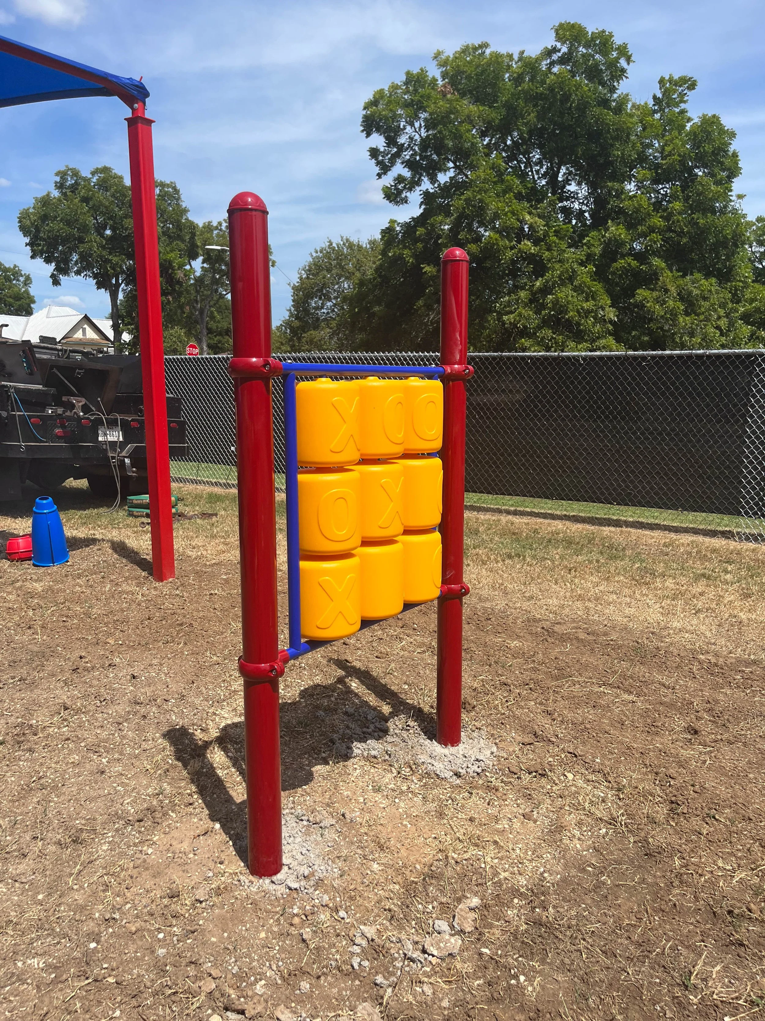 A colorful outdoor tic-tac-toe game with yellow X and O blocks on red and blue poles at a playground, with trees, a chain-link fence, and a clear blue sky in the background.