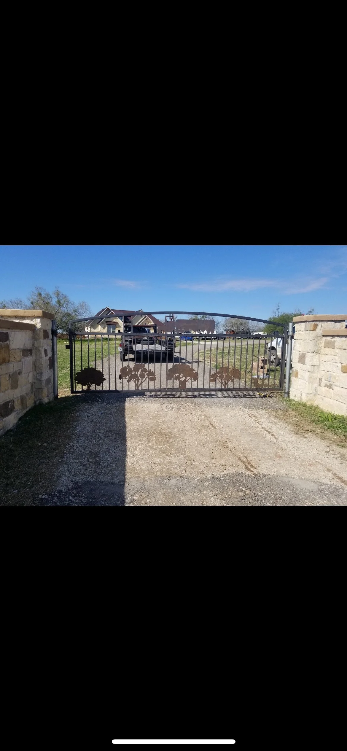 Black metal driveway gate with tree silhouette designs, supported by stone pillars, leading to a gravel driveway with cars and houses in the background under a clear blue sky.
