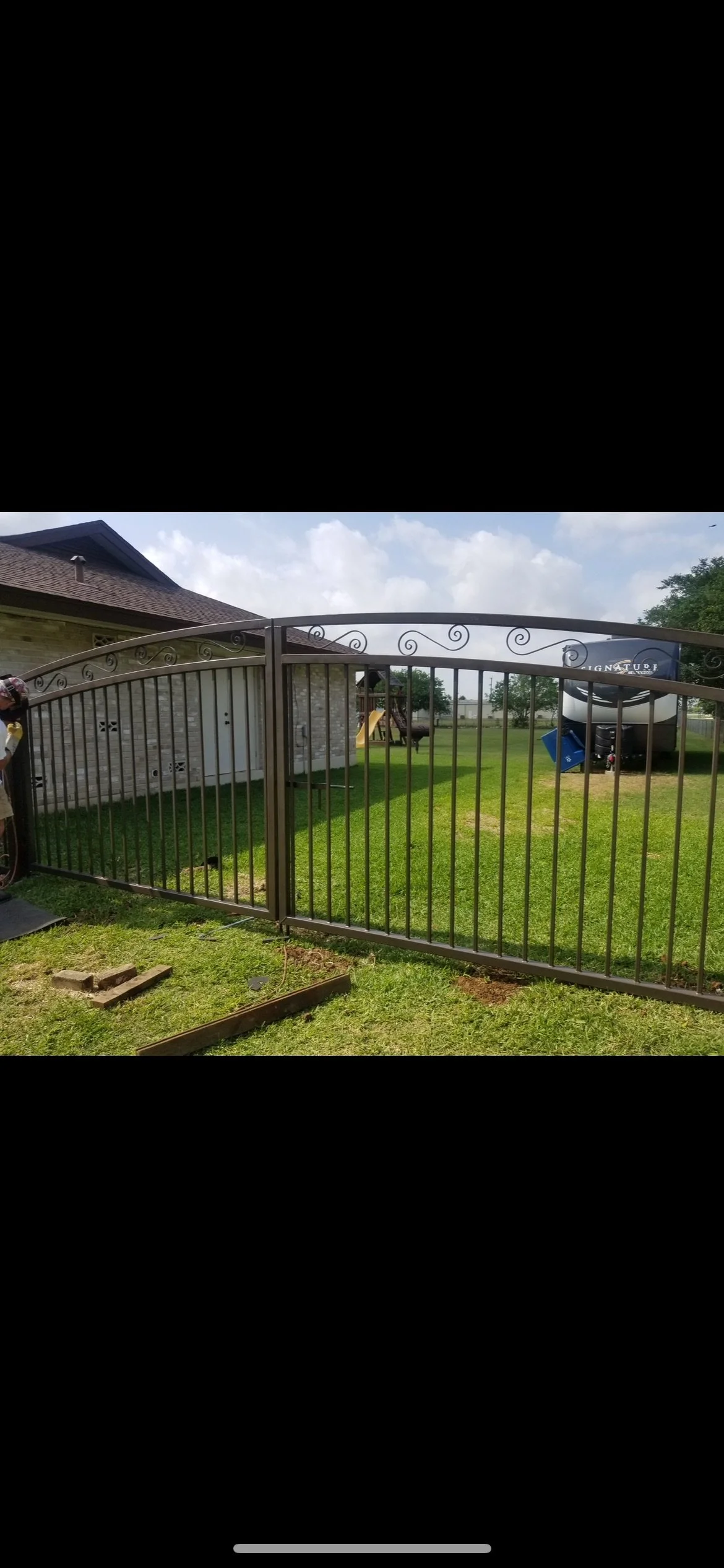 A backyard with a metal gate, grass, a brick house, a playground set with a slide, and a large black and white RV parked on the grass.