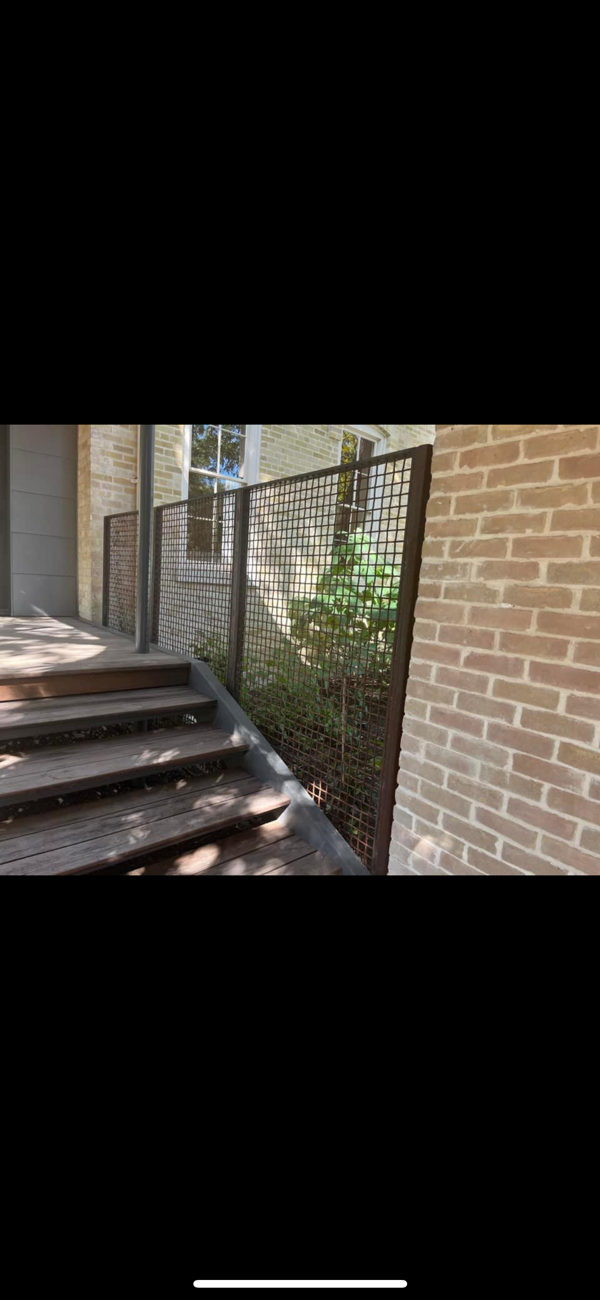 Wooden stairs leading up to a house with a brick exterior, black metal railing, and greenery along the side.