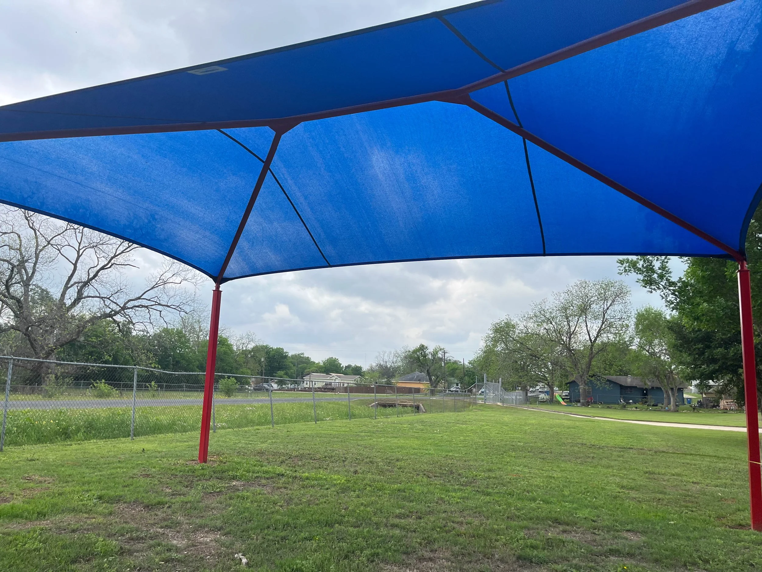 A large blue shade canopy supported by red poles over a grassy park area with trees, a chain-link fence, and some houses in the background, under a cloudy sky.
