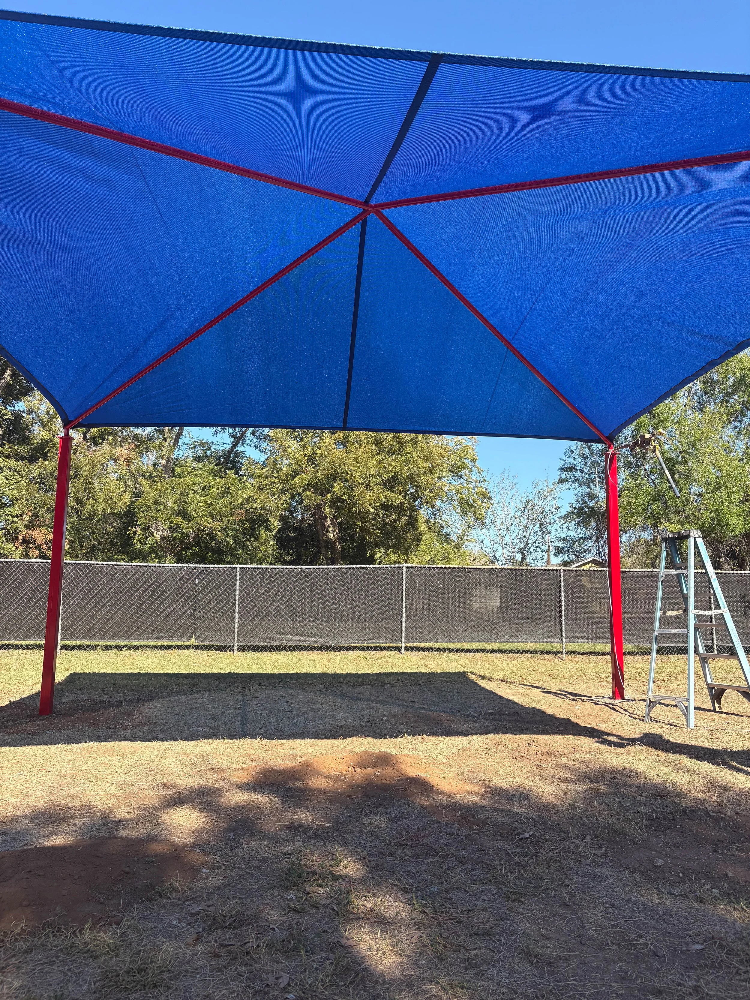 A blue shade structure with red poles on a grassy area, with a chain-link fence and trees in the background under a clear blue sky.