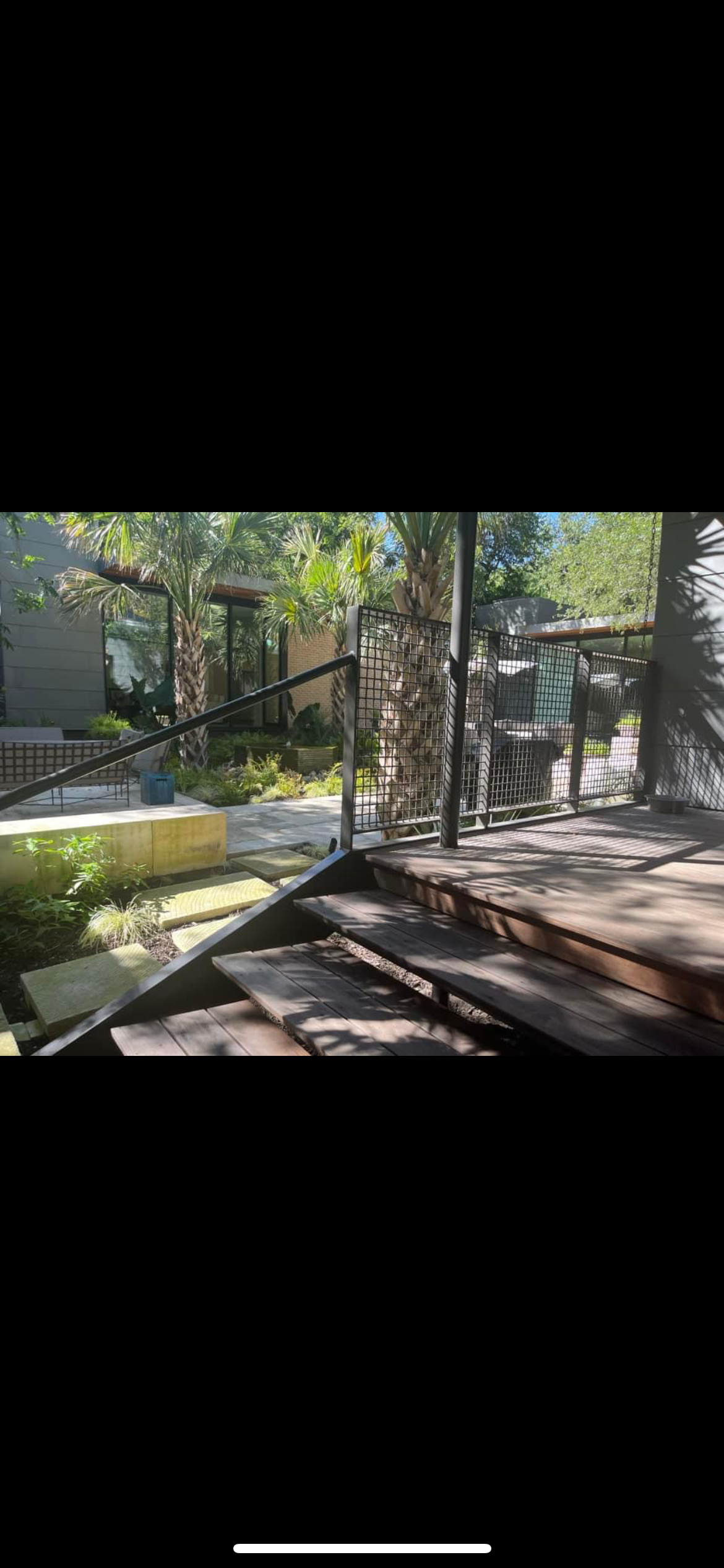 Outdoor patio area with wooden stairs, a metal railing, lush green plants, and tall palm trees in a modern residential backyard.