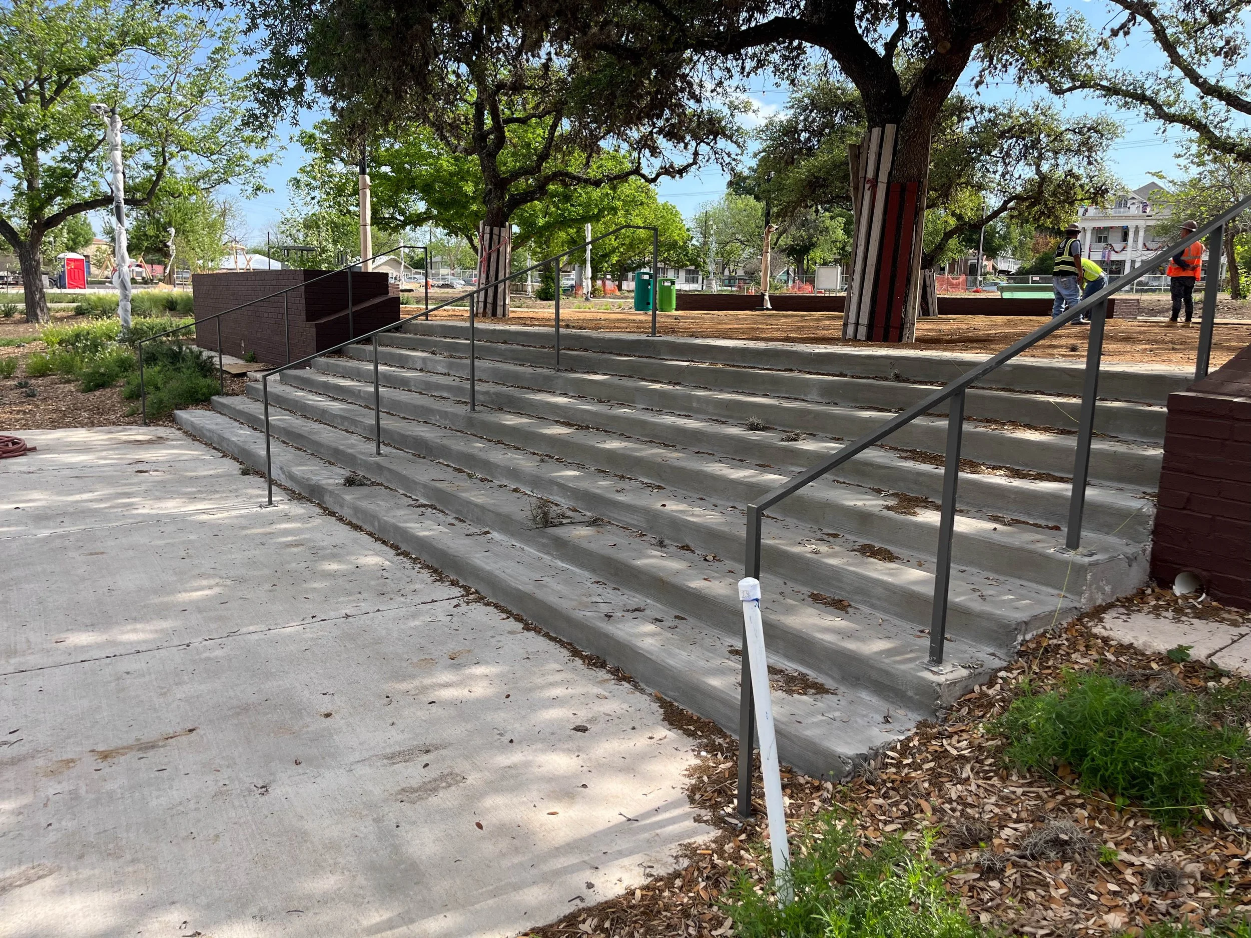 People working on a landscaping project in a park surrounded by trees, with a set of stairs, a railing, and trash bins visible.