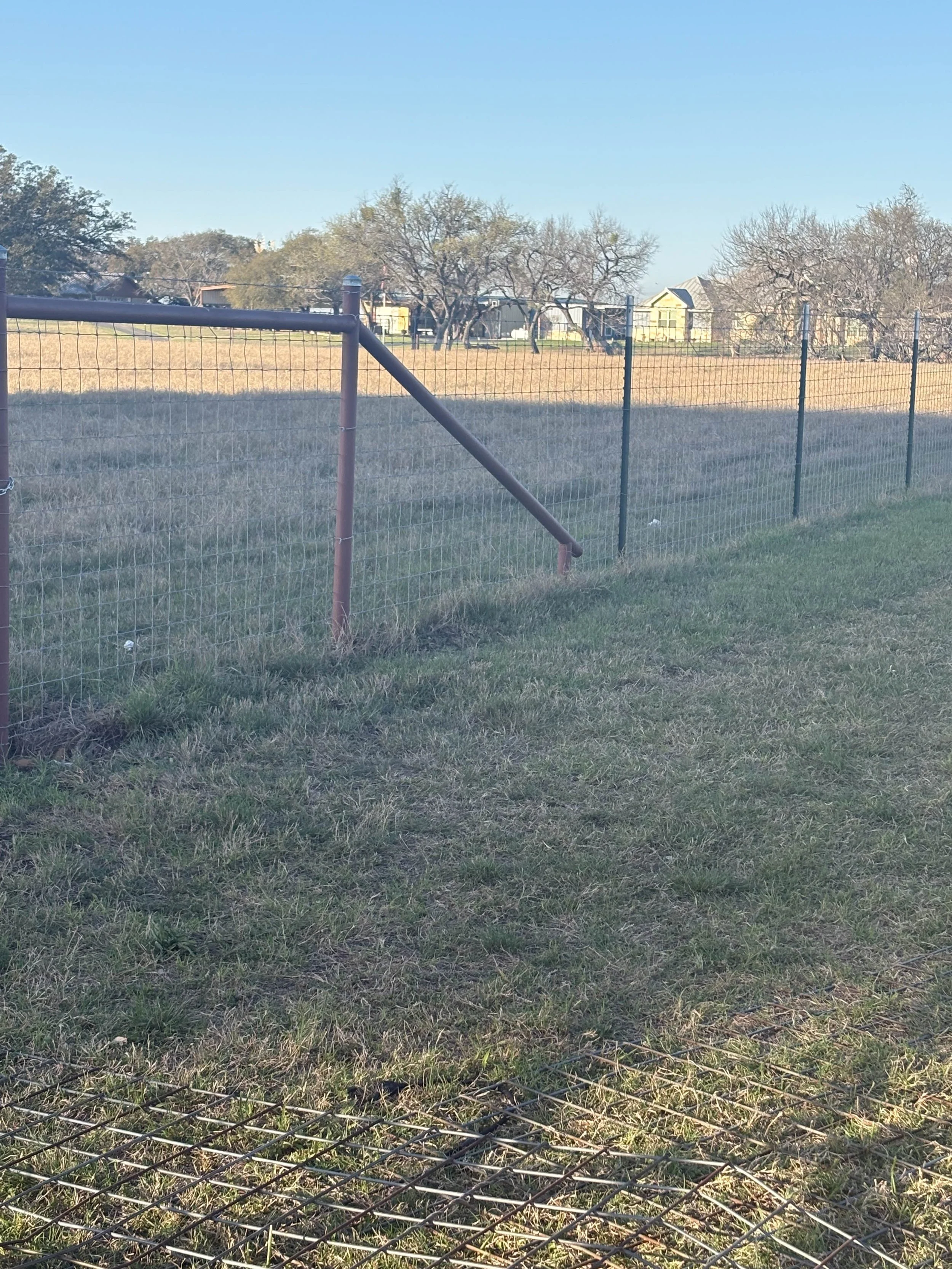 A wire fence with a metal frame on a grassy field, with houses and leafless trees in the background under a clear blue sky.