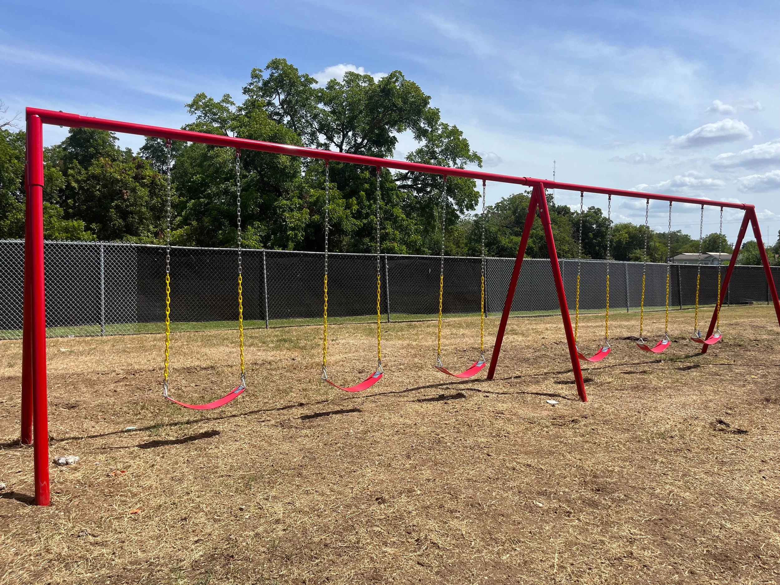 Red metal swing set with three swings, situated on a grassy area enclosed by a chain-link fence. There are trees and a blue sky with clouds in the background.