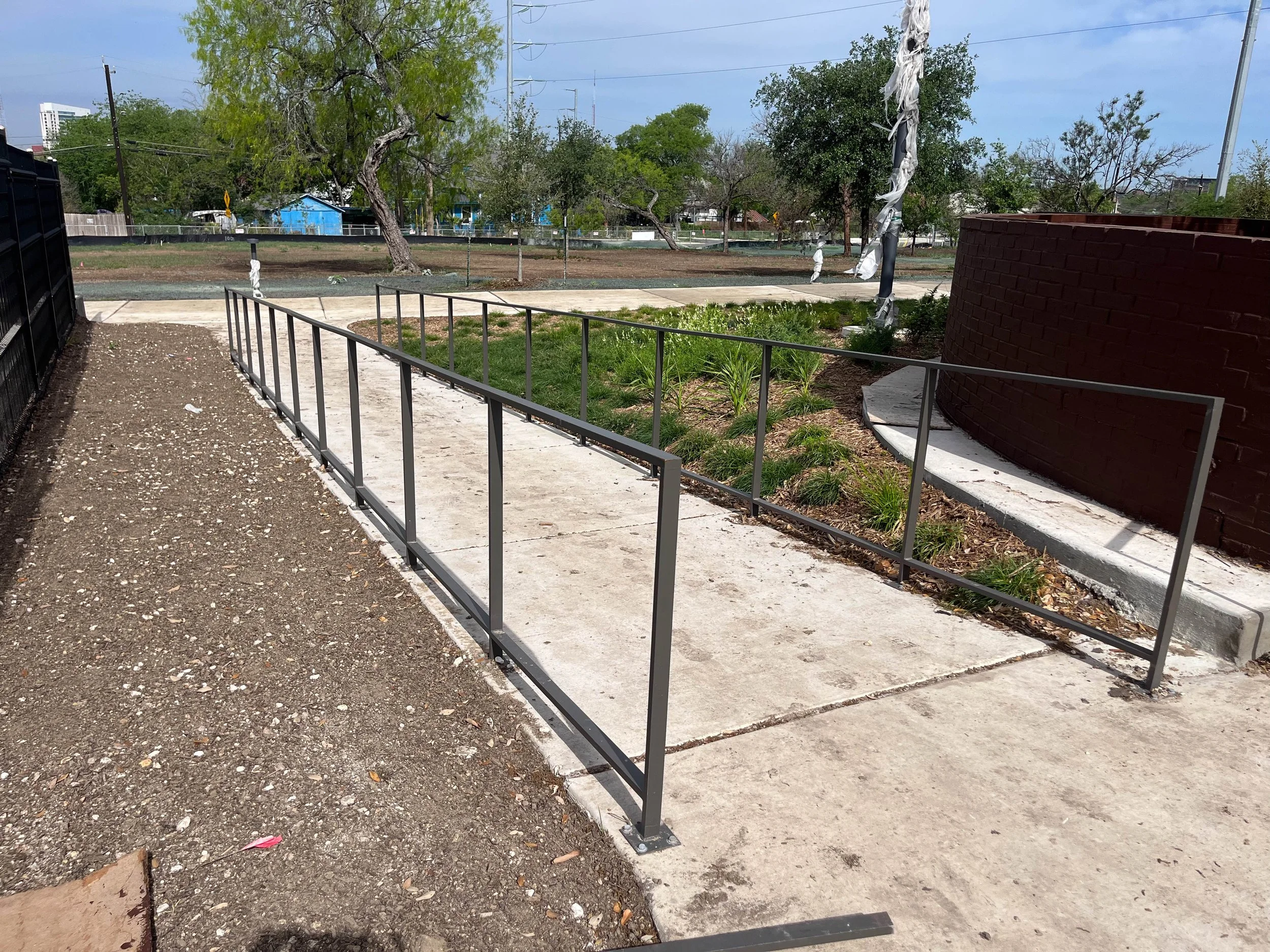A sidewalk with a metal railing next to a small patch of grass and plants, with two trees and a red brick curved wall across the sidewalk. In the background, there is a park or empty lot with grass and trees, some covered in white wrapping, and build