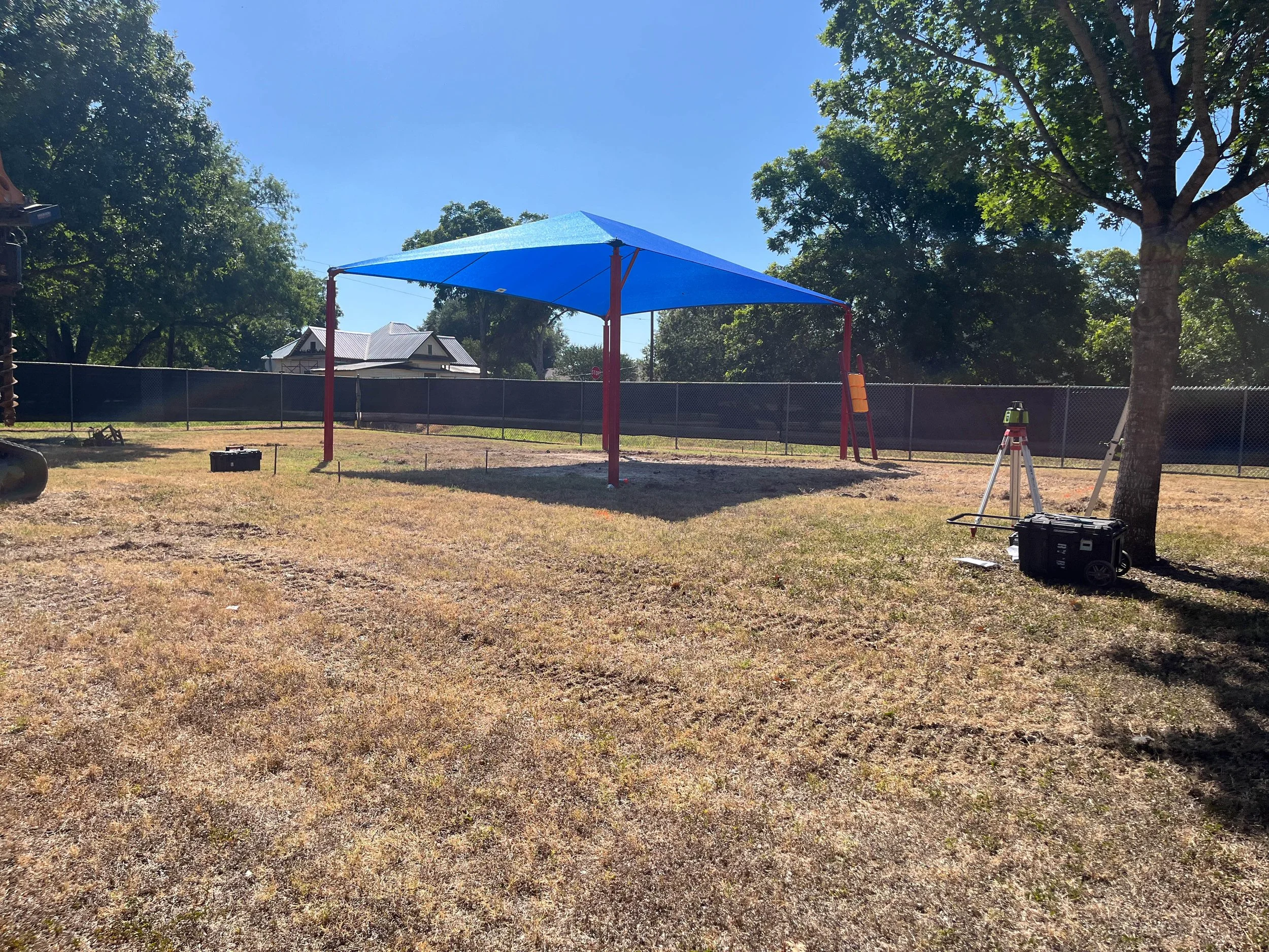 A backyard with a large blue shade sail, a tree, playground equipment, and a house in the background under a clear blue sky.
